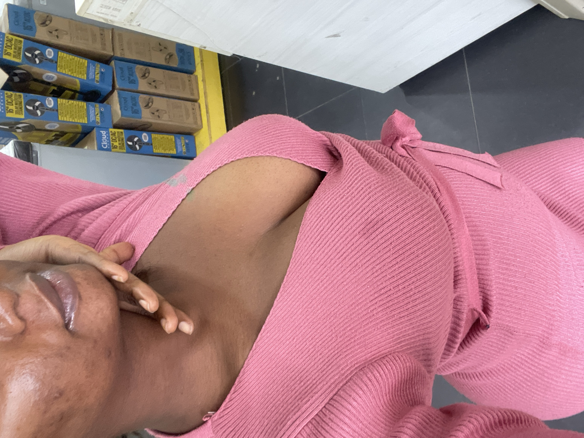 The image shows a close-up view of a person wearing a pink ribbed dress with a V-neckline. The person's hand is touching their chin. In the background, there are stacks of blue and brown boxes, some of which appear to be for standing fans, labeled "Cloud 16" DC/AC". The setting seems to be a retail environment, possibly a store in Ikeja, Nigeria, given the context. The lighting is moderate, and the focus is on the person and their attire.