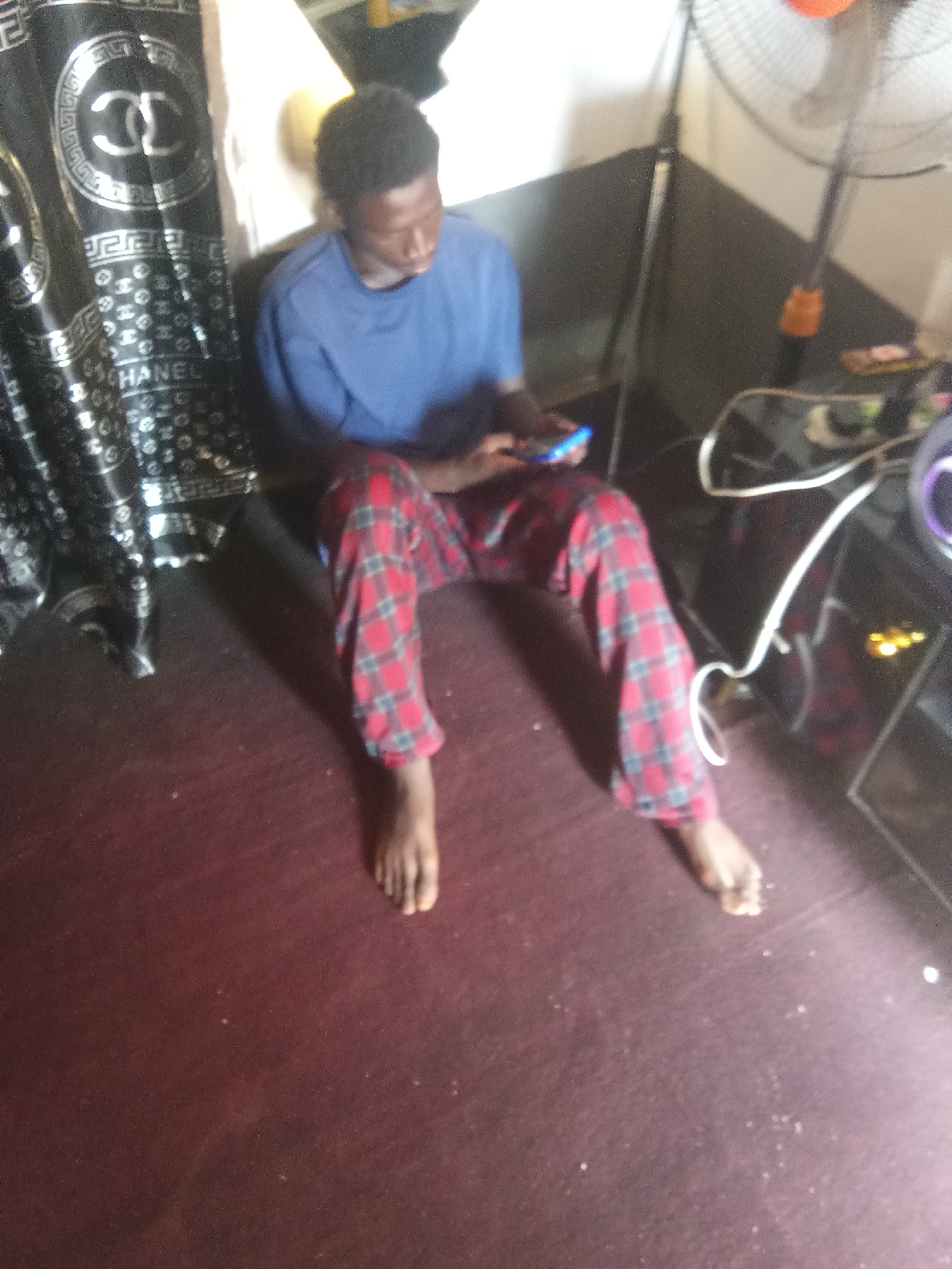 In a dimly lit room, a young Black male sits cross-legged on a dark red carpet. He is wearing a blue t-shirt and red and black checkered pajama pants. His attention is focused on a mobile phone in his hands. To his left, a black curtain with a silver "CC" logo and "CHANEL" lettering is partially visible. In the background, a standing fan with an orange base is visible. The environment appears to be an indoor residential space, possibly a bedroom or living area. The overall impression is one of casual relaxation and engagement with technology. The scene is consistent with a domestic setting that could be found in any urban area, including Jos, Nigeria.