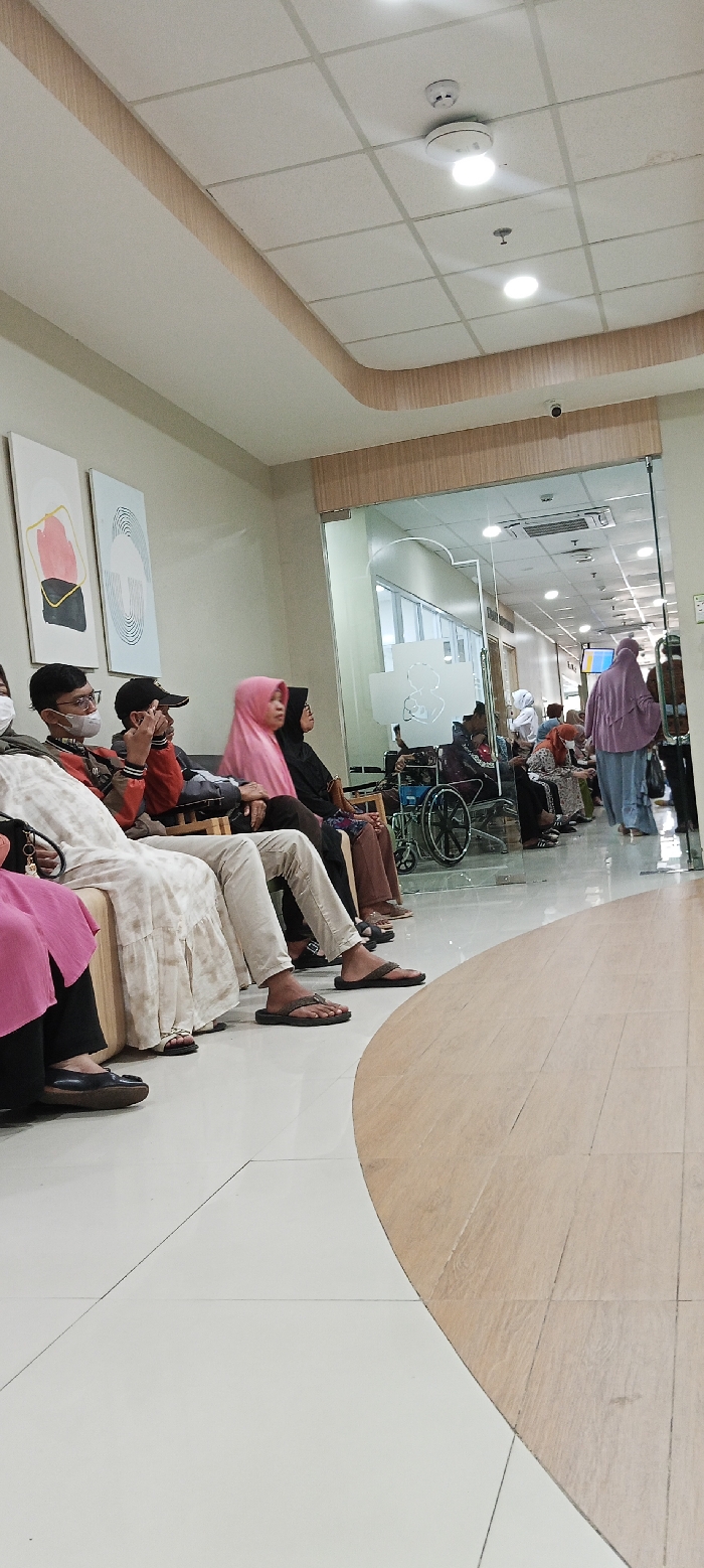 The image shows a waiting area in a hospital in Soreang, Indonesia. The waiting area has white walls and a white tiled floor. It is partially visible in the picture, and the rest of the waiting area can be seen through a large glass window. In the waiting area, a group of people sit on white seats, waiting. Most are wearing traditional clothing and face masks. There are some people in the waiting area, who are not fully visible, and they appear to be waiting as well. Behind the waiting area is a hallway that has beige tiles, with people walking through it. The people in the hallway are not fully visible. There is also a wheelchair partially visible in the photo, which is also not fully visible. There is an air conditioning vent and a security camera on the ceiling. The air conditioner is partially visible and the security camera is not fully visible. There are two paintings on the wall, one is of a pink square with a yellow border, and the other is of a white and yellow swirl pattern. The waiting area is clean and well-lit. It is a typical waiting area in a hospital.