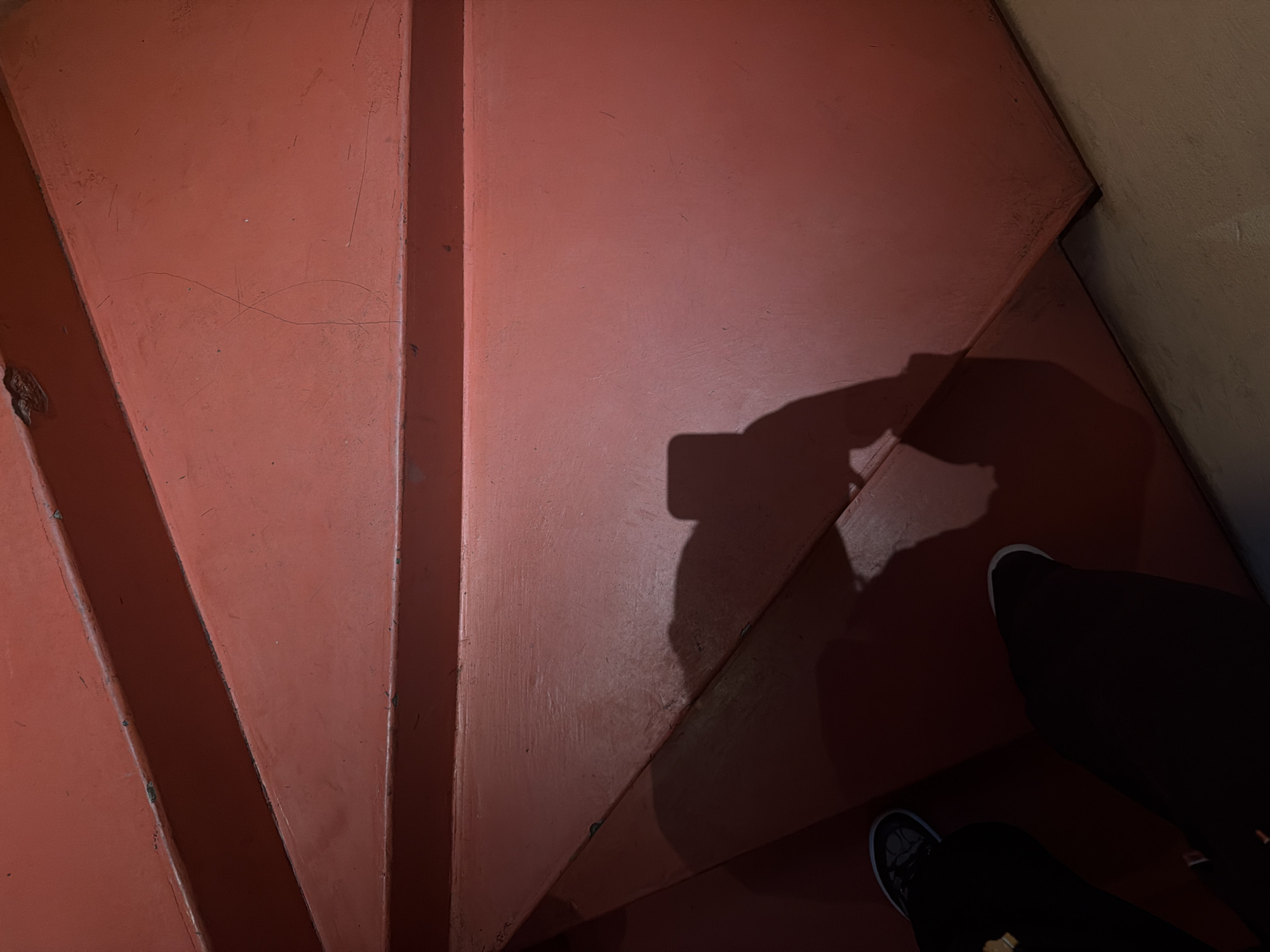 The image presents a top-down view of an interior staircase, likely within a building in Yerevan, Armenia. The stairs are finished in a faded reddish-orange paint, exhibiting widespread scratches, scuffs, and minor hairline cracks on the tread surfaces, indicating significant use. Darker wear patterns are apparent along the step edges. In the lower-left, the feet and lower legs of an individual are visible, wearing dark trousers and dark athletic shoes with light-colored soles/trims; one shoe displays a circular design. A prominent shadow, identifiable as a person holding a smartphone, is cast across the central and right portions of the staircase, suggesting the photographer's action and the direction of overhead illumination. The surrounding environment consists solely of the staircase and a small section of a lighter-colored floor at the bottom right. The precise location within Yerevan is not identifiable from the visual evidence presented.