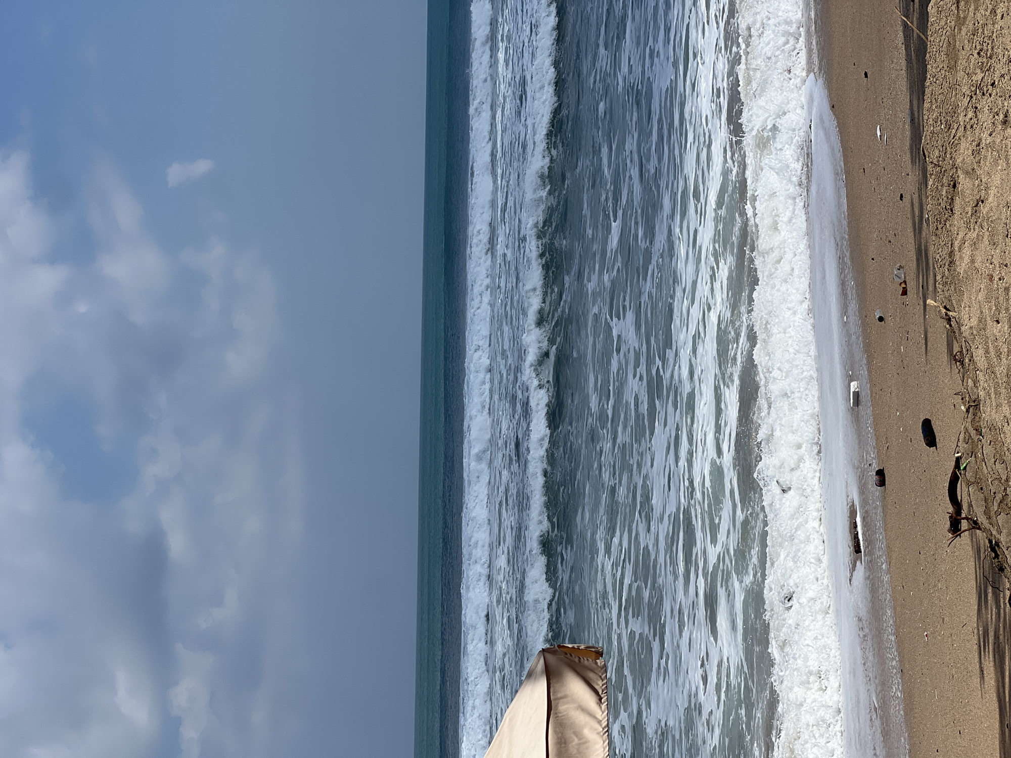 The image depicts a beach scene in Kuta, Indonesia. In the foreground, a sandy beach is visible, littered with various pieces of debris, including plastic bottles, a dark cylindrical object, and a piece of driftwood. The sand transitions to the ocean in the middle ground, where waves are breaking, creating white foam. The water is a deep blue-green color. In the far background, the ocean stretches to the horizon, meeting a pale blue sky dotted with scattered clouds. A portion of a beige beach umbrella is visible on the left side of the image, partially obscuring the view of the water. The overall impression is of a somewhat polluted beach with ongoing wave activity.