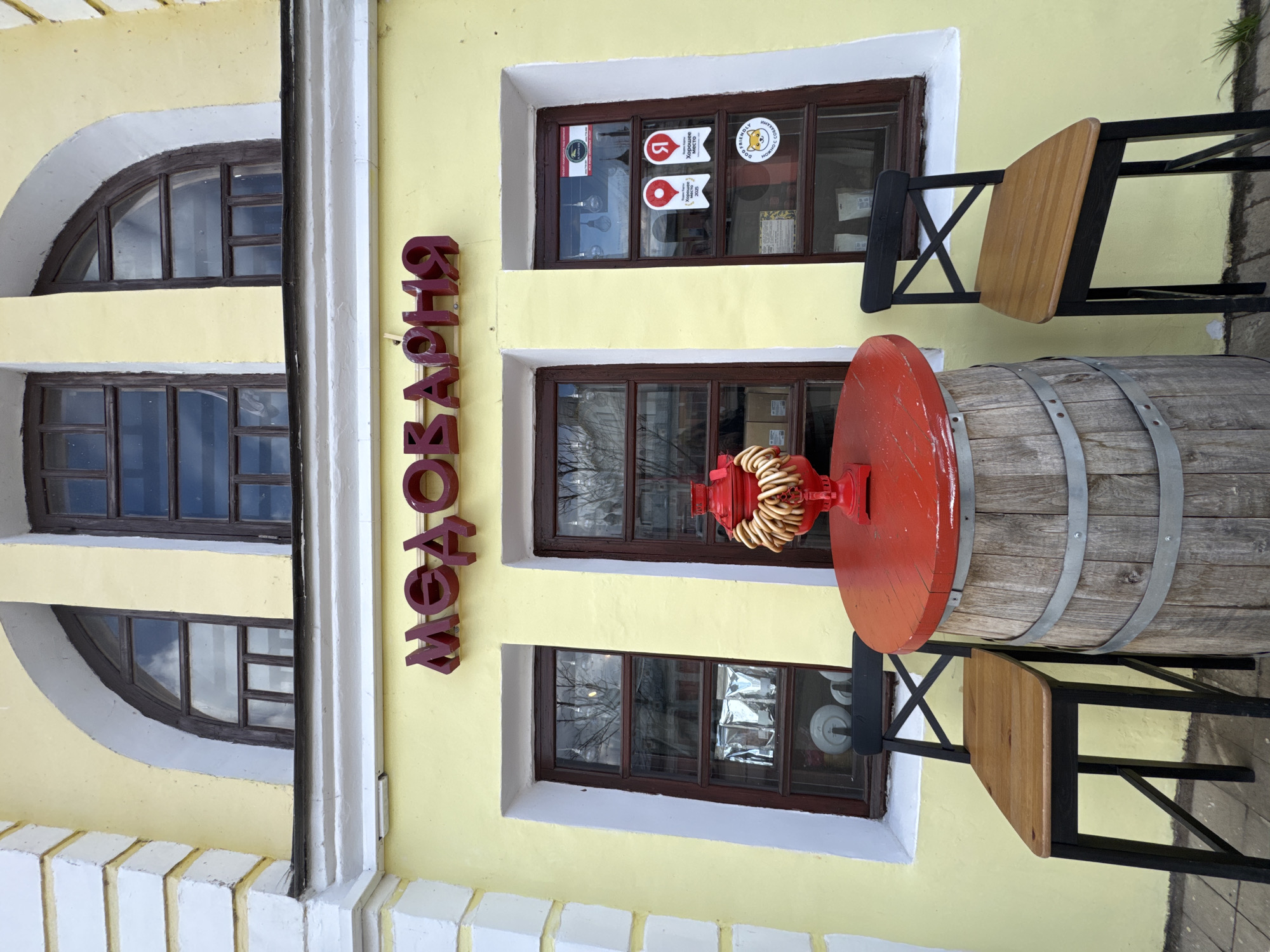 The scene depicts the exterior of a building, likely a commercial establishment, in Ново-Никольское, Russia. The building's facade is painted a light yellow, with white trim around the windows. There are three windows visible, two rectangular and one with an arched top. The building has a sign in Cyrillic script reading "Овощная". Outside, a barrel with a red tabletop and a cluster of pretzels on top serves as a table. Two wooden chairs are positioned on either side of the barrel table. Stickers are visible on the windows including ones that say "Dog Friendly".