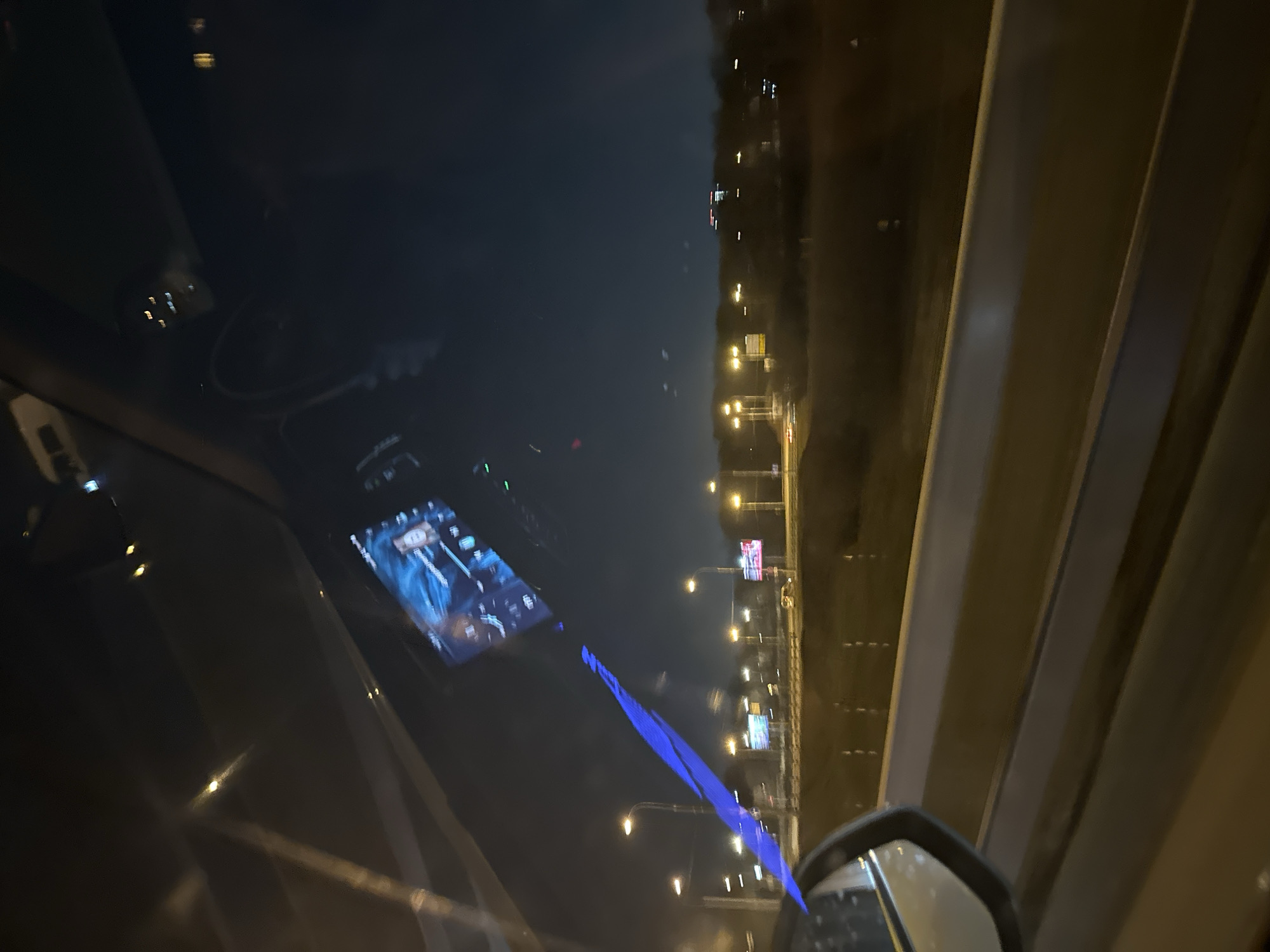 The image, taken at night from inside a vehicle, appears to show the vehicle driving on a highway or elevated road in Moscow, Russia. Streetlights line the roadway, and highway signs are visible in the distance. Inside the vehicle, a navigation system is mounted on the dashboard, displaying a map. The dashboard reflects the ambient light from the city, as well as the reflection from the screen of the navigation system. The side mirror of the car is also visible in the lower right corner of the image.