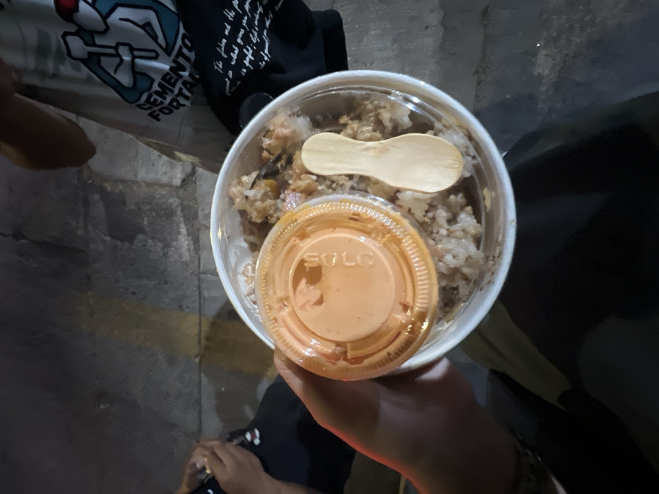 An overhead, dimly lit image captures a person's hand holding a clear plastic food container in León, Mexico. The container holds a mixed rice dish, topped with a small plastic cup featuring an orange lid labeled "SOLO" and a small wooden spoon. To the left, the arm and lap of a seated individual wearing dark pants and a white shirt are visible. To the right, another person's torso is seen, wearing a light-colored t-shirt with a blue and white graphic depicting two figures holding tools, and the text "CEMENTO FORTAL". The ground beneath is a dark, textured paved surface, possibly a sidewalk, with a faint yellow marking. The scene appears to be taking place at night or in a low-light indoor setting.