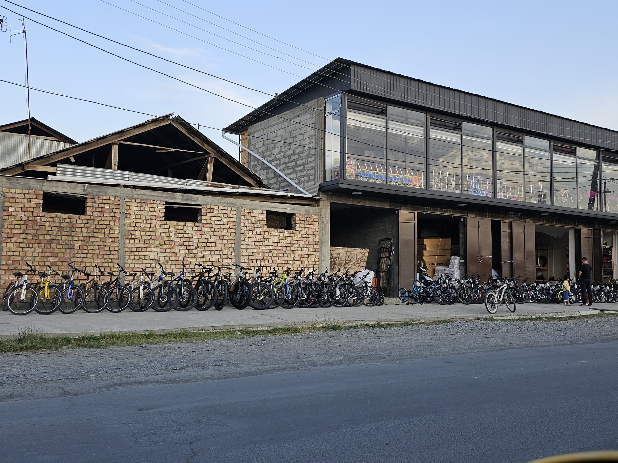 The image depicts a row of bicycles parked in front of a building in the city of Özgön, Kyrgyzstan. The building appears to be a commercial establishment, possibly a shop or a warehouse, with a modern upper level featuring large glass windows. Adjacent to this, on the left, is an older structure made of brick with a partially constructed roof. The bicycles are lined up along the sidewalk, with some standing upright and others leaning against each other. A few individuals are visible near the entrance of the building, one of whom is standing and looking at the bicycles. The scene suggests a retail or rental service for bicycles.
