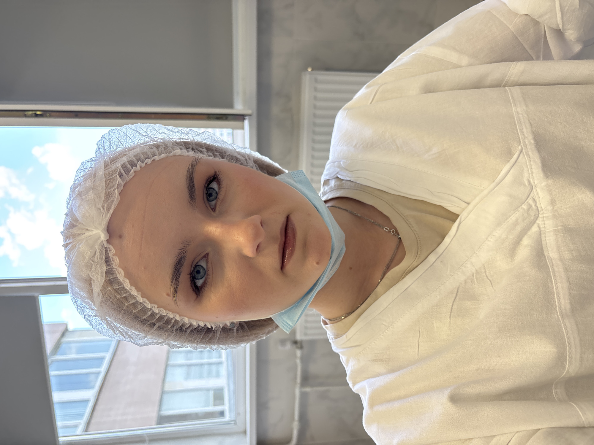 An upright-oriented shot features a young woman looking directly at the viewer, likely in a professional or clinical setting in Saint Petersburg, Russia. She has fair skin, blue eyes, and a neutral expression with slightly parted lips. Her light hair is covered by a disposable white bouffant cap, and a light blue surgical mask is pulled down, resting under her chin.
She is wearing a crisp white medical gown or lab coat over a light-colored, possibly beige, top. A thin silver chain necklace is visible around her neck.
The background on the left side of the image (when upright) reveals an indoor wall, possibly concrete or tiled, with a white radiator, suggesting an interior space. The right side of the background shows a window with a clear view of a bright blue sky with white clouds, indicating daytime. Below the sky, reflections or partial views of modern buildings are discernible, hinting at an urban environment outside. The overall lighting is bright and natural. There is no visible text in the image.