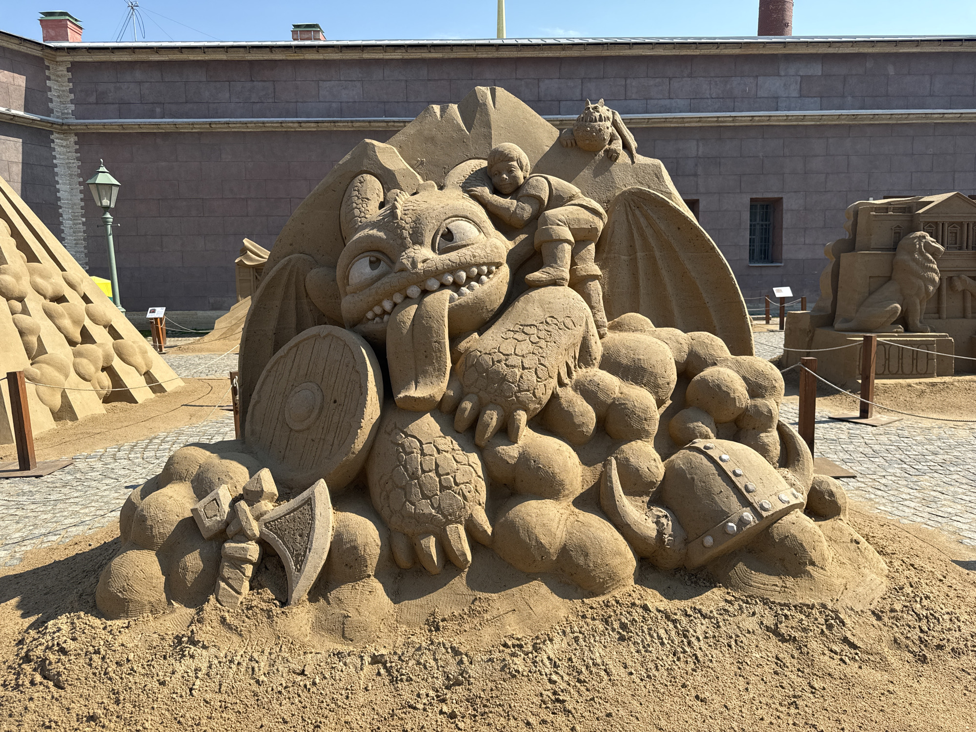 The image shows a sand sculpture in Saint Petersburg, Russia. The sculpture depicts characters and objects from the "How to Train Your Dragon" franchise. A boy figure is leaning on the head of a dragon. The sand sculpture also incorporates a shield, axe, and helmet. In the background is a building with a reddish hue and rectangular window openings. Cobblestone paving surrounds the sculpture. Other sand sculptures can be seen in the background.