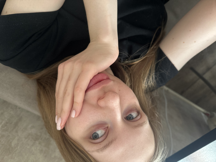 A close-up, angled photograph, taken in Chaikovsky, Russia, shows a fair-skinned individual, identified as female, with long light-colored hair. Her blue eyes are directed upwards and to the left, and her lips are slightly parted. Her right hand, exhibiting light pink nail polish, rests against her right cheek and jawline. She is wearing a black short-sleeved t-shirt. The background on the left features a dark floor, a light-colored structural element, and the underside of a dark surface. The right side of the background displays a textured, light brown upholstered material.