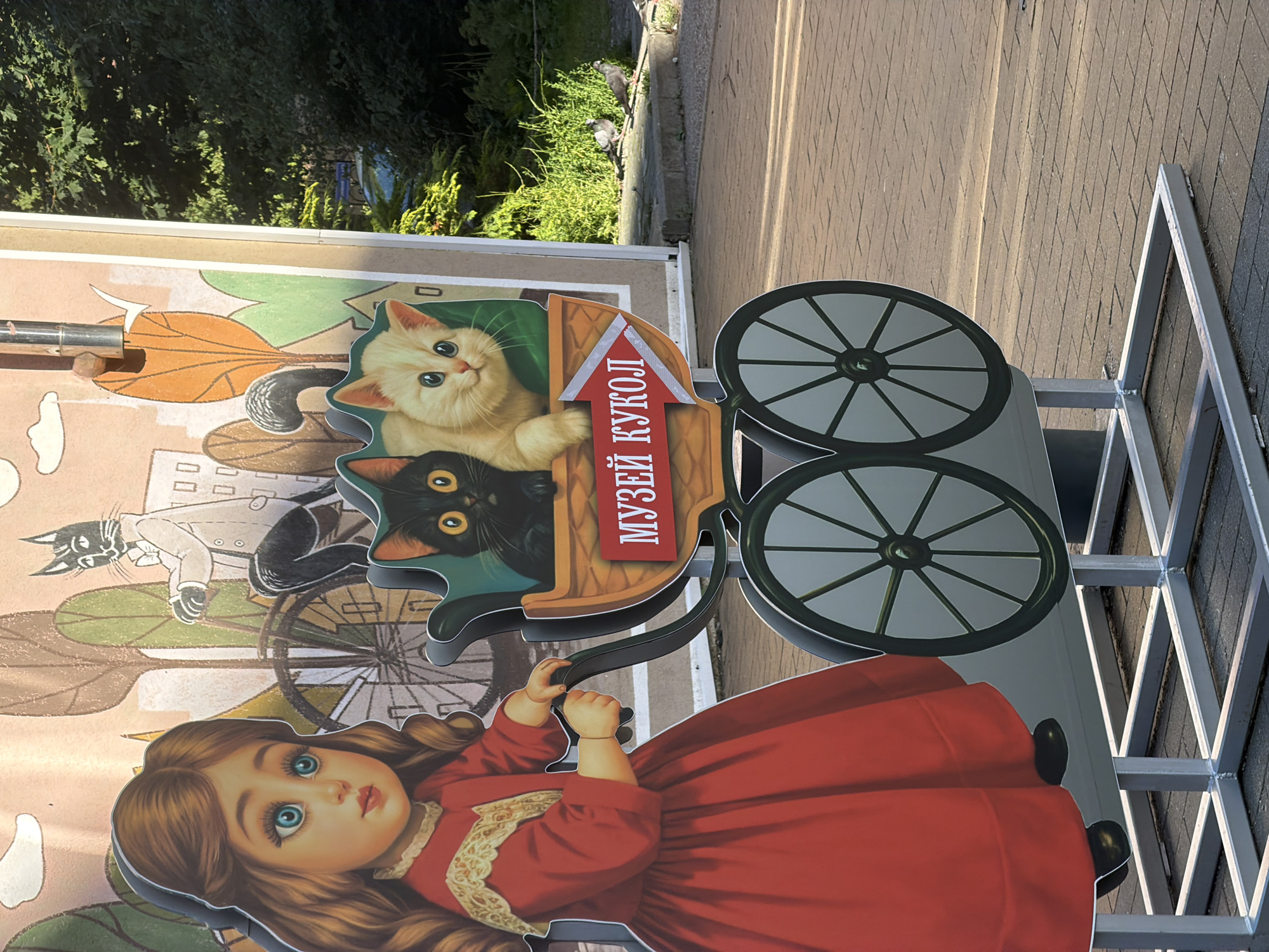 The image depicts a life-sized cutout display of a little girl in a red dress, pushing a stroller containing two kittens. The backdrop features a stylized illustration of a city scene with trees and a cat riding a bicycle. The text on the stroller reads "МУЗЕЙ КУКОЛ" (Doll Museum). The display is situated on a paved walkway, with lush green trees and foliage visible in the background, bathed in sunlight. The overall scene suggests an outdoor exhibit or promotional display, likely in a public space. This cutout is located in Zelenogradsk, Russia, a city known for its feline population and cat-themed attractions, including the "Cat Museum."