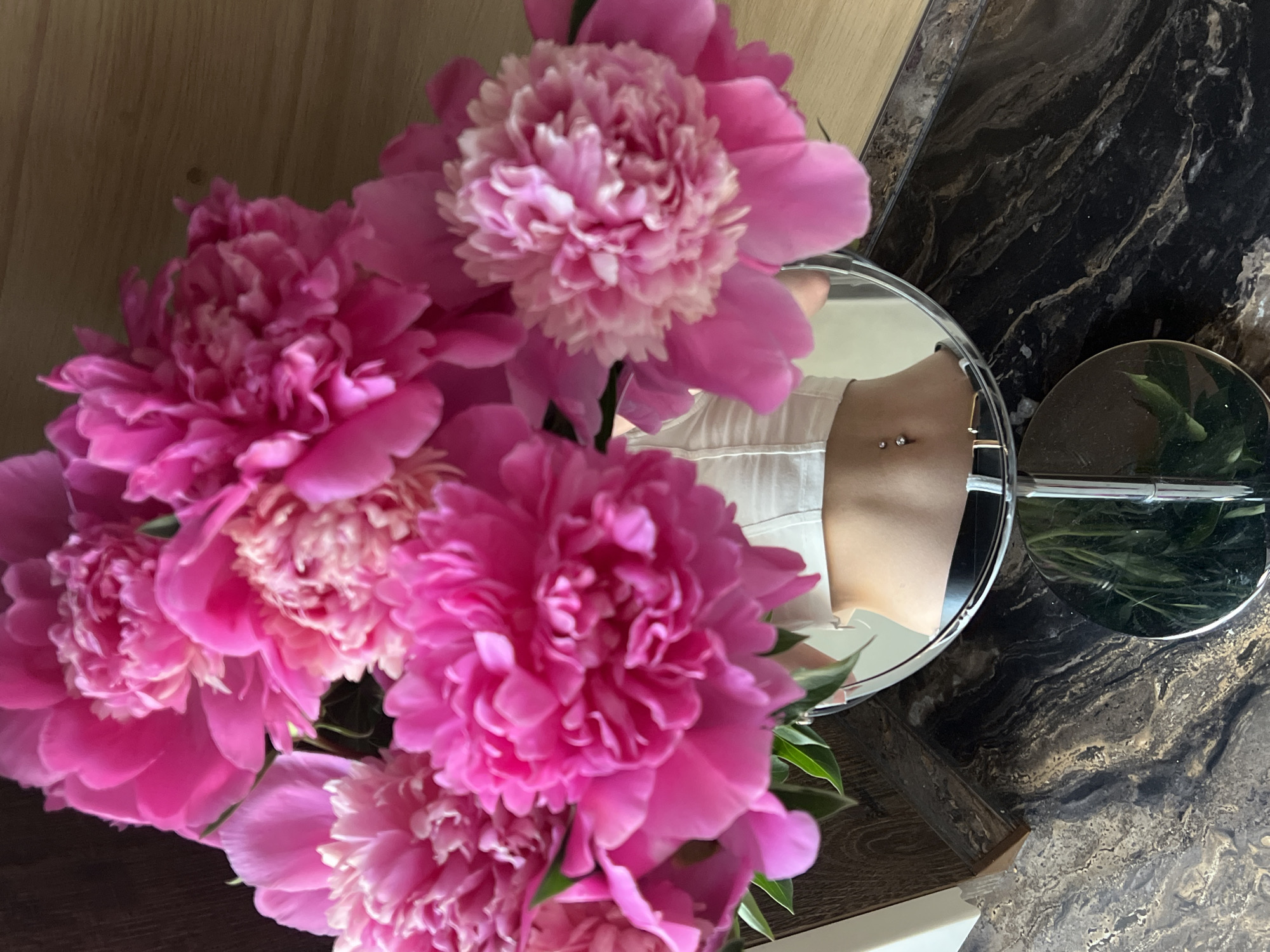 A bouquet of bright pink peonies is arranged in a clear glass vase. The vase sits on a dark, marbled surface. In the reflection of a circular mirror placed in front of the vase, a person's midriff is visible, wearing a white, cropped top and a black belt with a gold buckle. The person has a navel piercing. The background consists of a wooden surface. The image was taken in Ternopil, Ukraine.