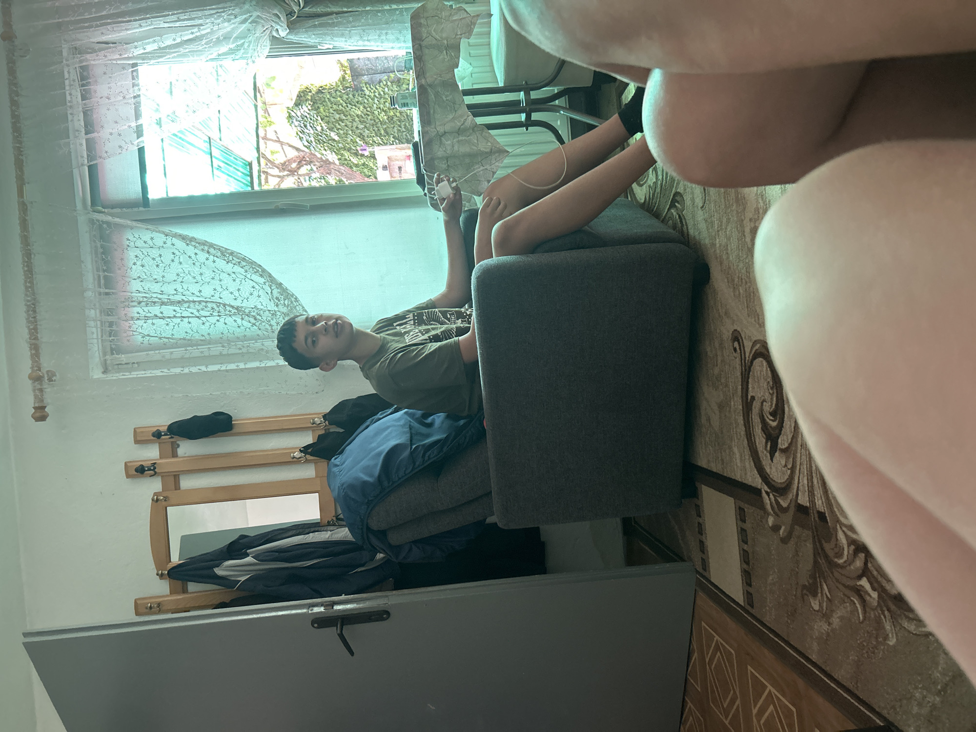 In Govedare, Bulgaria, a young male with short, dark hair and a light complexion is seated in a gray armchair. He is wearing a dark olive green t-shirt with a graphic print. His right hand rests on a white object, possibly a phone charger, with a white cable extending from it. The armchair is positioned in front of a window draped with white, patterned lace curtains. Outside the window, lush green ivy covers a wall, and there is a glimpse of a green roof structure. To the left of the armchair, a wooden coat rack with several coats and hats hangs on the wall. A closed gray door is visible on the far left of the image. The floor is covered with a patterned rug. The foreground of the image is dominated by the blurred out legs of another person.