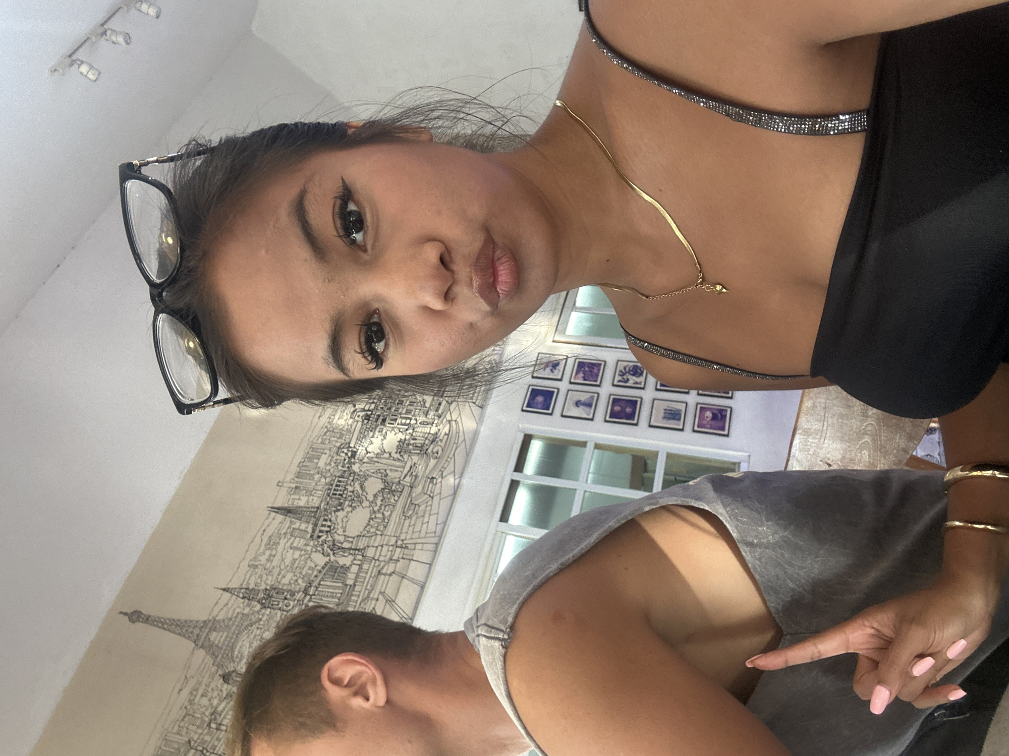 A photograph shows two people. One person is wearing glasses and a black top with sparkly straps and a gold necklace, and has an arm around another person who is wearing a gray shirt. The second person is pointing their finger. The background shows a wall with a drawing of a city and a window with pictures on the wall. Based on the drawing of the Eiffel tower in the cityscape, the drawing is of Paris. However, the location in the city of Sukawati, Indonesia is unsubstantiated.