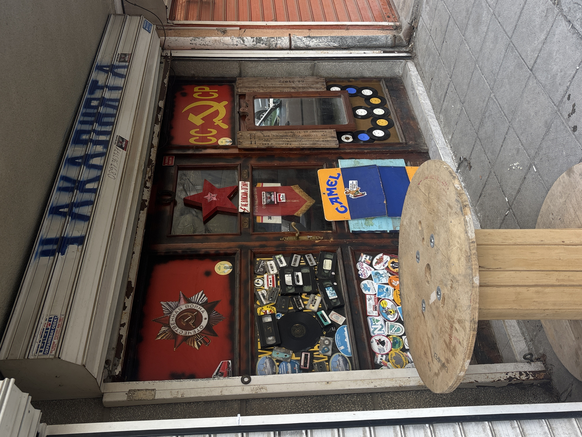 The image shows the storefront of a shop called "Chakalniata" (ЧАКАЛНЯТА) in Varna, Bulgaria. The storefront is cluttered with Soviet-era memorabilia and various objects. The shop's sign is painted in blue on the closed metal shutter. The shop's display window is divided into six panes, each containing different items: Soviet symbols (hammer and sickle), vinyl records, a mirror, a red star, a Camel cigarette ad, cassette tapes, a war medal, and various stickers. A large wooden spool lies in front of the shop's window, partially obscuring the view. The street pavement is gray and speckled.