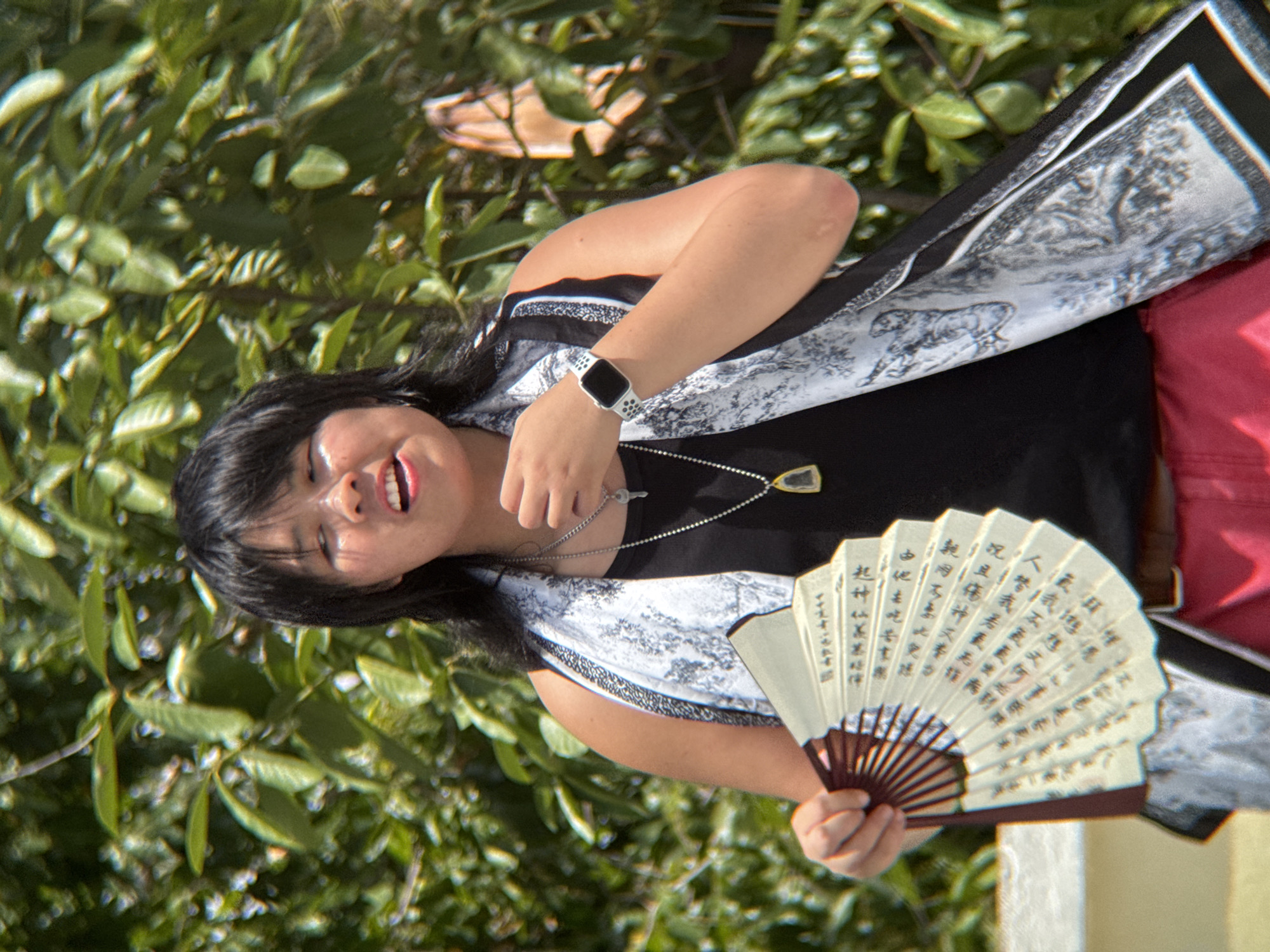 A young Asian woman stands outdoors, holding a white folding fan with Chinese characters written on it. She is wearing a black tank top with a white and black patterned scarf draped around her shoulders, red pants, and a silver smartwatch on her left wrist. Her mouth is open as if she is speaking or singing. The background is filled with lush green foliage and bright sunlight, suggesting a warm, sunny day. The scene appears to be from a park or garden in Nha Trang, Vietnam.
