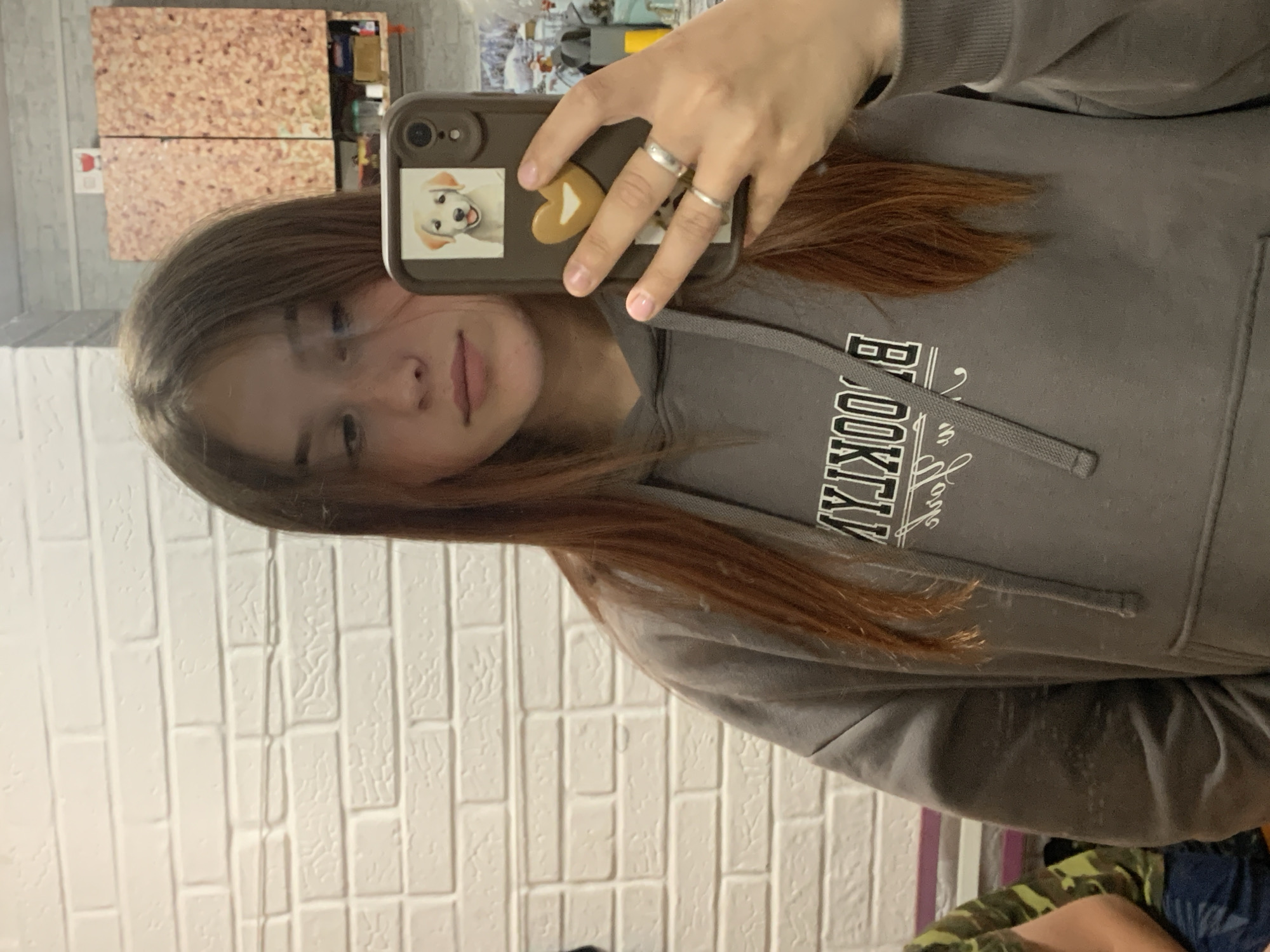 A person is taking a selfie in front of a white brick wall, wearing a gray hoodie with "BROOKLYN" printed on it. The person is holding a phone with a brown case adorned with a sticker of a dog and a heart. The location is not discernible from the image; it is not possible to verify if the picture was taken in Фоки, Russia.