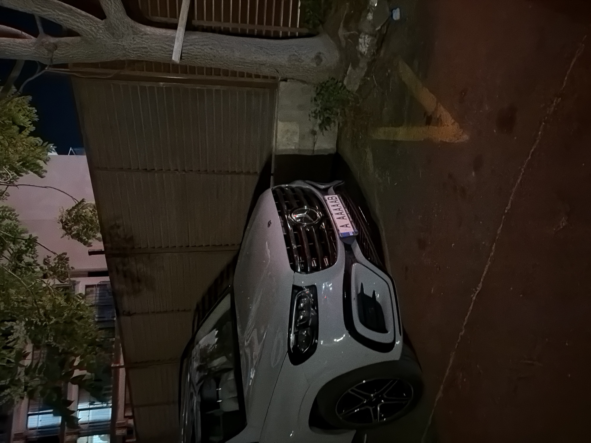 A light-colored Mercedes-Benz GLS SUV is parked in an outdoor parking spot next to a wooden fence and a tree. The car has its front facing the viewer, and the license plate reads "A AAAAAB". The scene appears to be at night or in low-light conditions, with artificial lights visible in the background. The parking spot is marked with yellow lines on the ground.