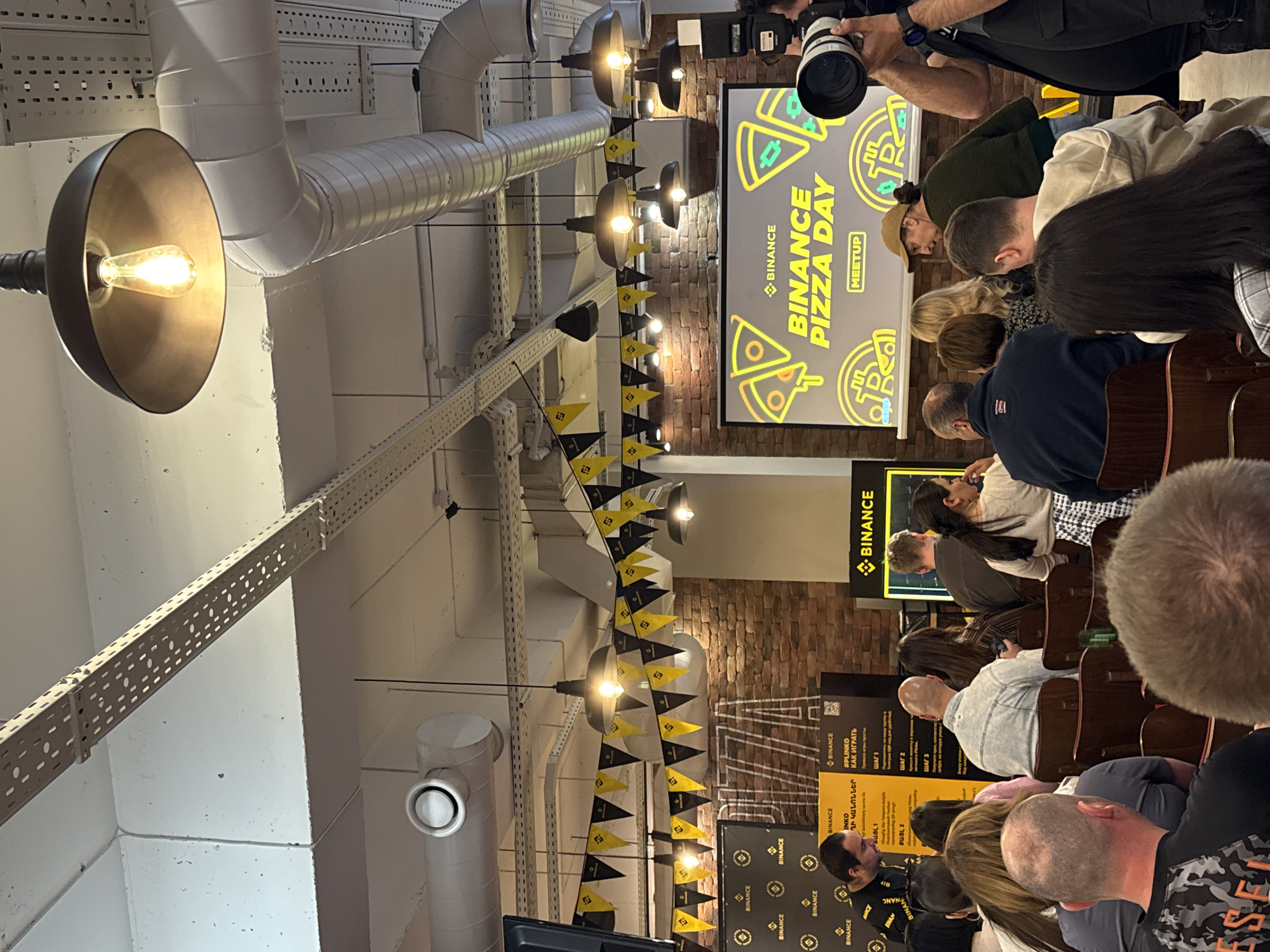 The image captures a Binance Pizza Day meetup, likely in Yerevan, Armenia, given the presence of Armenian text on promotional materials. Attendees are seated, facing a screen displaying the Binance logo and the event name. Overhead, industrial-style lighting fixtures hang from a white ceiling, with a string of black and yellow pennants. The crowd is diverse, with varying hairstyles and clothing. A photographer with a professional camera is positioned near the screen, documenting the event. The room appears to have exposed brick walls and a casual, industrial aesthetic.