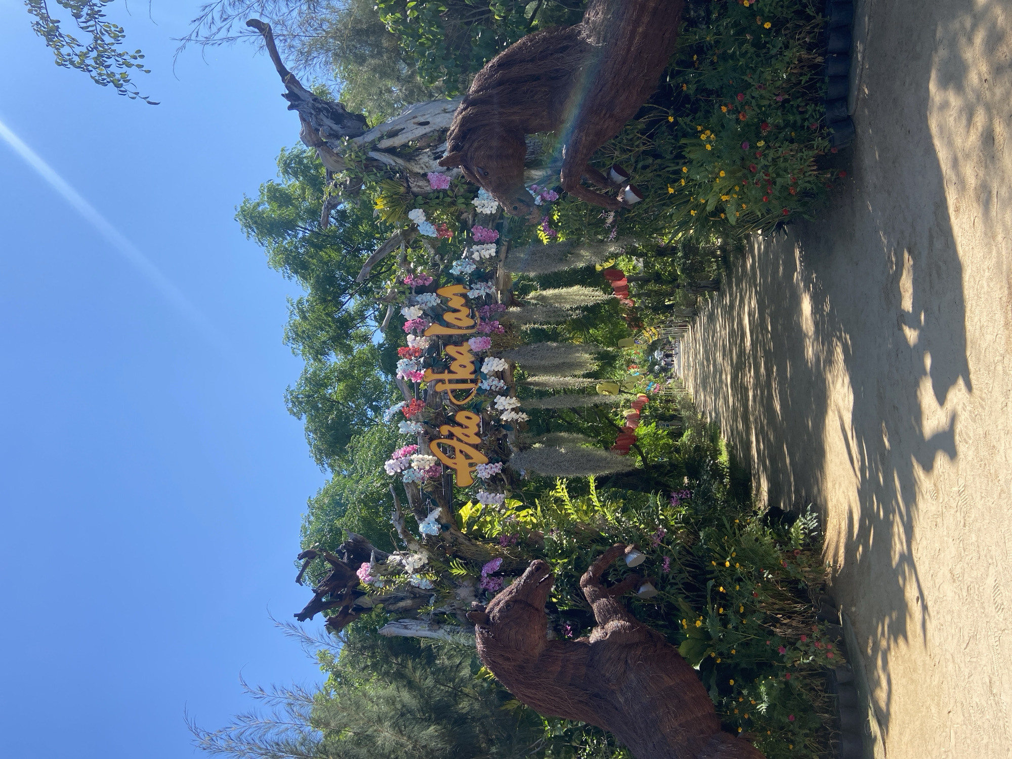 The image depicts an entrance to a park or garden area, possibly a tourist attraction, in Hoa Thang, Vietnam. The foreground is a sandy path lined with lush green foliage and colorful flowers. On either side of the path, at the entrance, are large, rustic sculptures of horses, crafted from natural materials like wood and vines. Above the path, an archway made of weathered wood is decorated with a vibrant array of artificial flowers in shades of pink, white, and blue. A yellow sign with Vietnamese text, "Đảo Hoa Lan," is prominently displayed on the archway, identifying the location as "Orchid Island." Hanging from the archway are strands of Spanish moss and what appear to be small, decorative lanterns. Further down the path, a few people can be seen walking, suggesting the area is open to visitors. The sky is a clear, bright blue, indicating a sunny day. The overall scene conveys a welcoming and whimsical atmosphere, characteristic of a tropical garden destination.