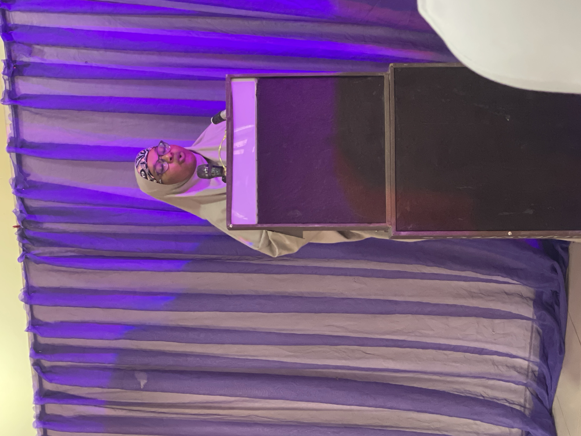 A person wearing a hijab and glasses is speaking into a microphone in front of a backdrop of purple and grey striped fabric. The person is positioned to the left of two large, dark rectangular objects, likely speakers or monitors. The backdrop is illuminated with purple lighting, creating a stage-like atmosphere. The person appears to be presenting or speaking at an event. The scene is indoors, with no indication of the time of day or weather conditions. There is no visible text in the image. The location context is Unguwar Shanu, Nigeria.