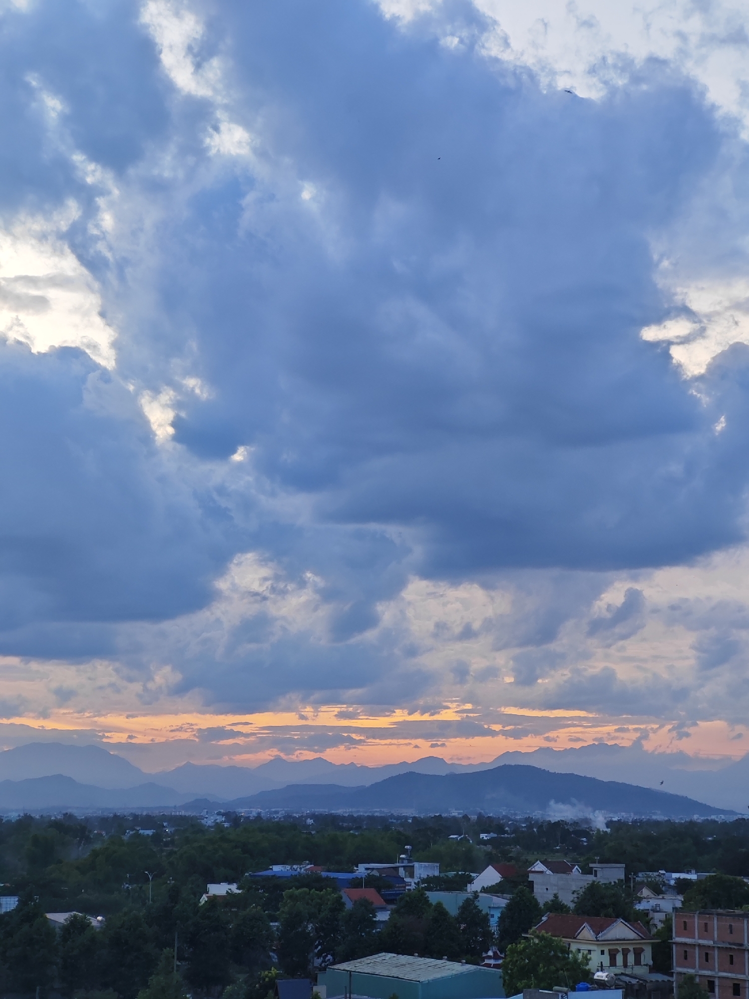 A wide-angle, vertical photograph captures a serene landscape from a high vantage point, likely overlooking the outskirts of Đà Nẵng, Vietnam, near sunset. The sky is dominated by voluminous, dark blue clouds that are partially illuminated by a soft, orange glow on the horizon, suggesting the sun is low in the sky or has just set. Amidst these clouds, two small, dark shapes that appear to be birds are visible, one in the upper right quadrant and another higher up and to the left.
Beneath the dramatic sky lies a sprawling cityscape transitioning into rural terrain. A range of majestic, blue-hued mountains forms the backdrop, their peaks softened by atmospheric haze. In the middle ground, a dense canopy of lush green trees covers rolling hills. Peeking through the foliage are various buildings, including houses with tiled roofs in shades of orange and white, and some with exposed brickwork. A few streetlights are discernible, casting faint illumination. A wispy trail of smoke rises from a point in the distance near the mountains, hinting at activity within the landscape. The overall impression is one of tranquility and natural beauty at dusk, with signs of human settlement integrated into the environment.