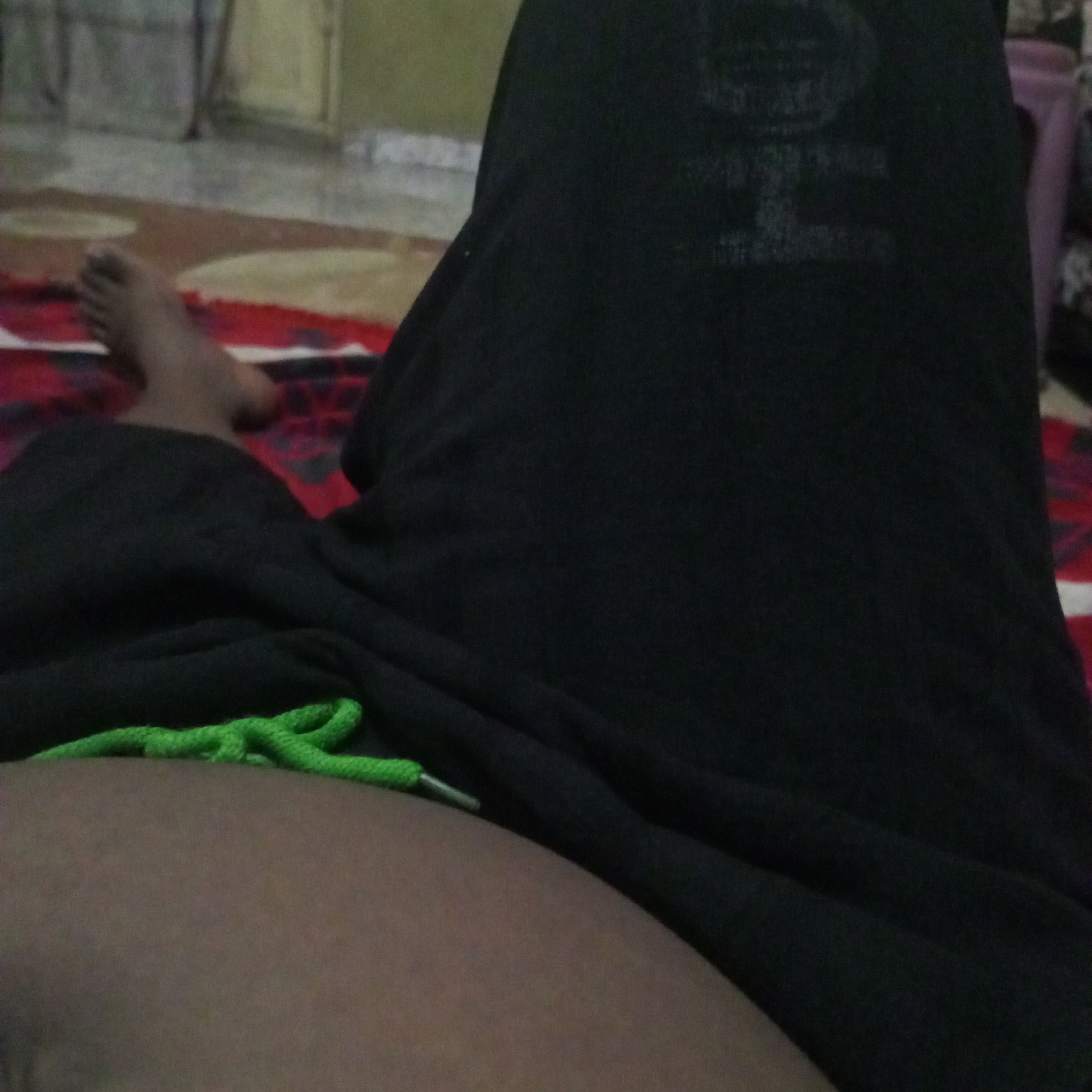 The image shows a close-up view of a person's legs and torso, covered by dark fabric, possibly clothing. A green drawstring is visible near the bottom left of the frame, attached to the dark fabric. The person appears to be lying down on a patterned rug, which has red and beige elements. In the background, out of focus, a wall and possibly furniture are dimly visible. The scene is indoors, suggesting a domestic environment. The exact location within Howayi, Nigeria cannot be determined from the visual information provided.