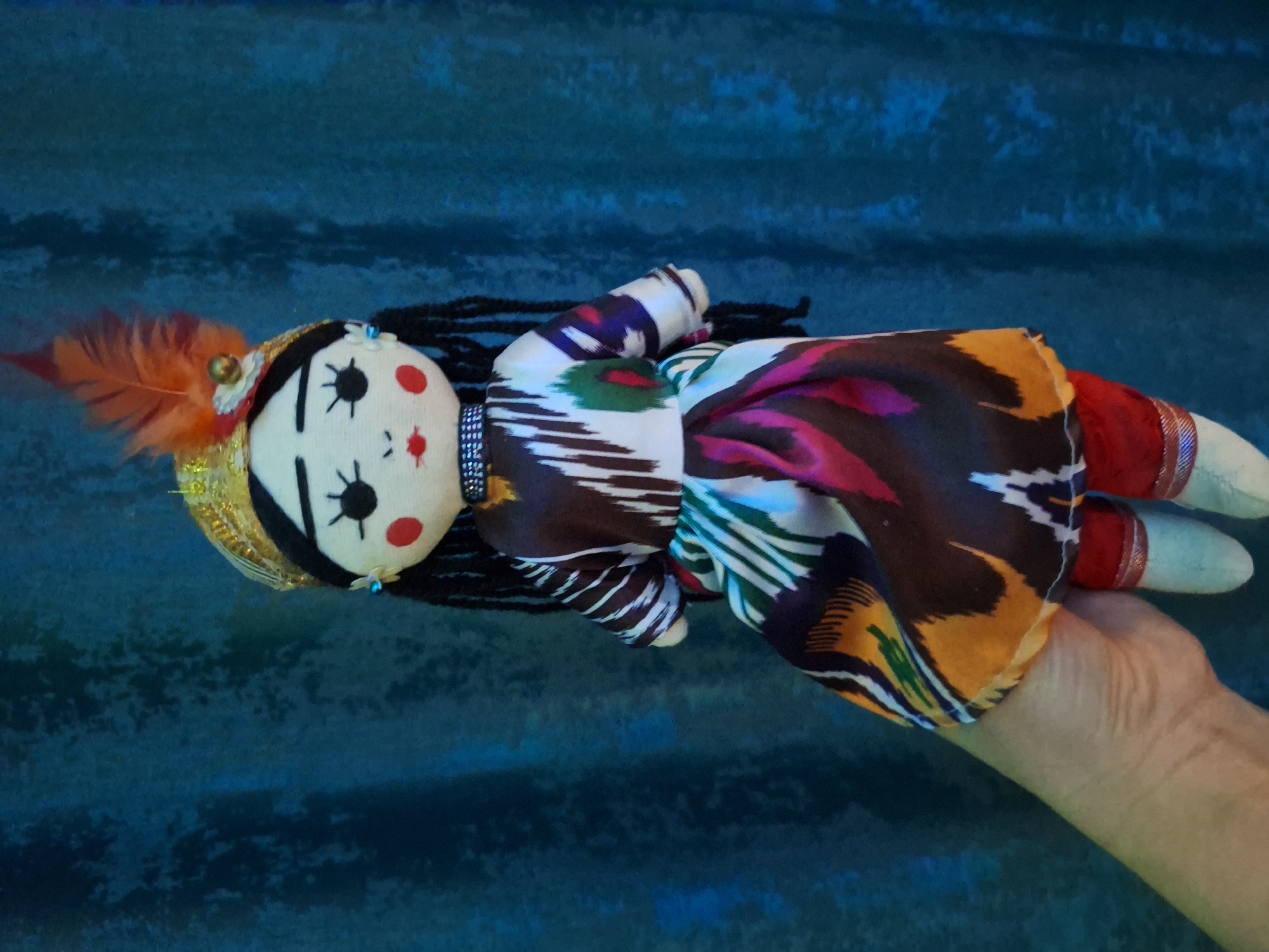 The image displays a vibrant, handmade doll held by a person's hand against a textured, dark teal background. The doll features a white fabric face with simple black stitched eyes, radiating lashes, round red cheeks, and small red lips, framed by long black braided yarn hair.
A golden, sequined band adorns the doll's head, embellished with a prominent orange feather and a small golden bead. The doll is dressed in a brightly patterned fabric, possibly an ikat print, showcasing a rich combination of brown, white, purple, green, yellow, and red in an abstract, geometric design. Its fabric arms are covered in the same distinctive pattern, while one leg is visible with a red cuff leading to a white fabric foot.
A fair-skinned adult hand gently supports the doll, presenting it to the viewer. The background provides a consistent, deep blue or dark teal hue with a visible, uneven texture, resembling a fabric or painted wall, but offering no specific location cues. No text is visible in the image. The scene suggests an indoor setting, focusing on the intricate details of the handcrafted doll.