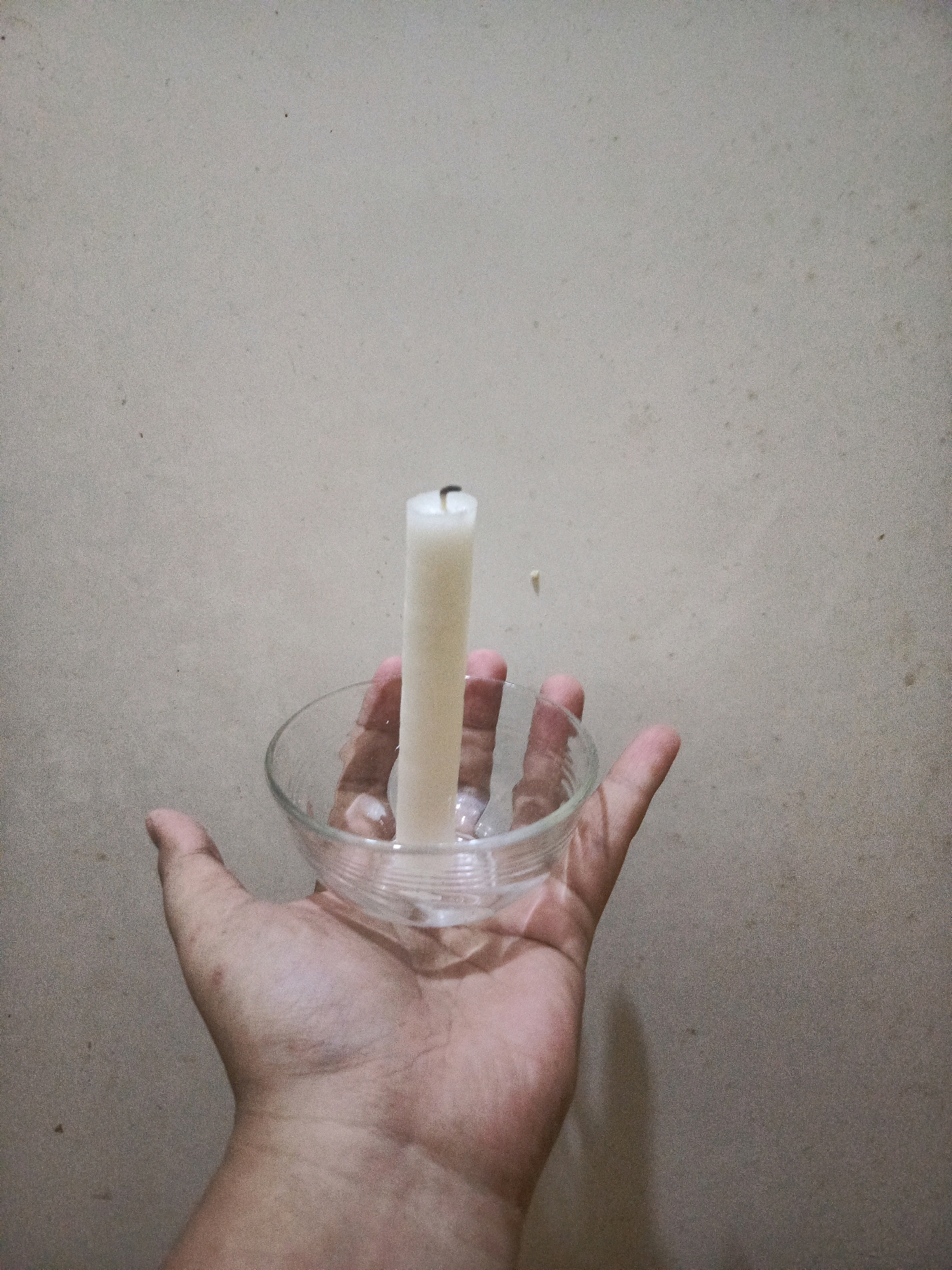 A close-up, waist-up shot shows a hand holding a clear glass bowl containing a white candle. The candle, appearing to be unlit, stands upright in the bowl. The background is a plain, textured wall, appearing to be a neutral beige color, with some subtle dirt or scuff marks. The lighting suggests an indoor environment, possibly with minimal natural light, as the scene is evenly lit and there are no strong shadows. The image composition focuses on the hand and the candle, with the background providing a simple backdrop. The scene is located in Balaraja, Indonesia.