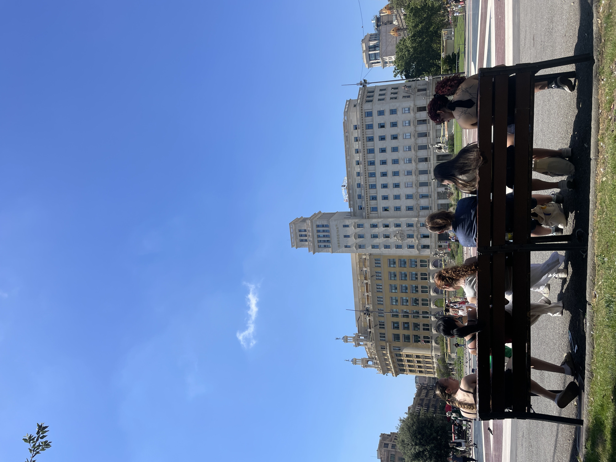 Six women are sitting on a dark brown park bench, viewed from behind, facing a large, ornate building in a city square under a clear blue sky. The building, which appears to be a significant architectural landmark, has a pale facade with many windows and decorative elements, including turrets. In the foreground, a strip of green grass borders the bench. The women are dressed casually in summer clothing. The woman on the far left wears a green top and white shorts, her legs crossed. The woman next to her has a dark top and dark shorts. The third woman from the left has a light-colored tank top and white pants. The fourth woman from the left wears a dark patterned top and dark shorts. The fifth woman has a light-colored top and appears to be wearing shorts. The woman on the far right has a dark tank top and dark shorts. Several large white shopping bags are placed on the ground in front of the bench. In the background, beyond the square, are more buildings and trees, and people are visible walking in the distance, suggesting a public, urban environment in Barcelona, Spain. The overall scene depicts a relaxed daytime gathering in a prominent city location.