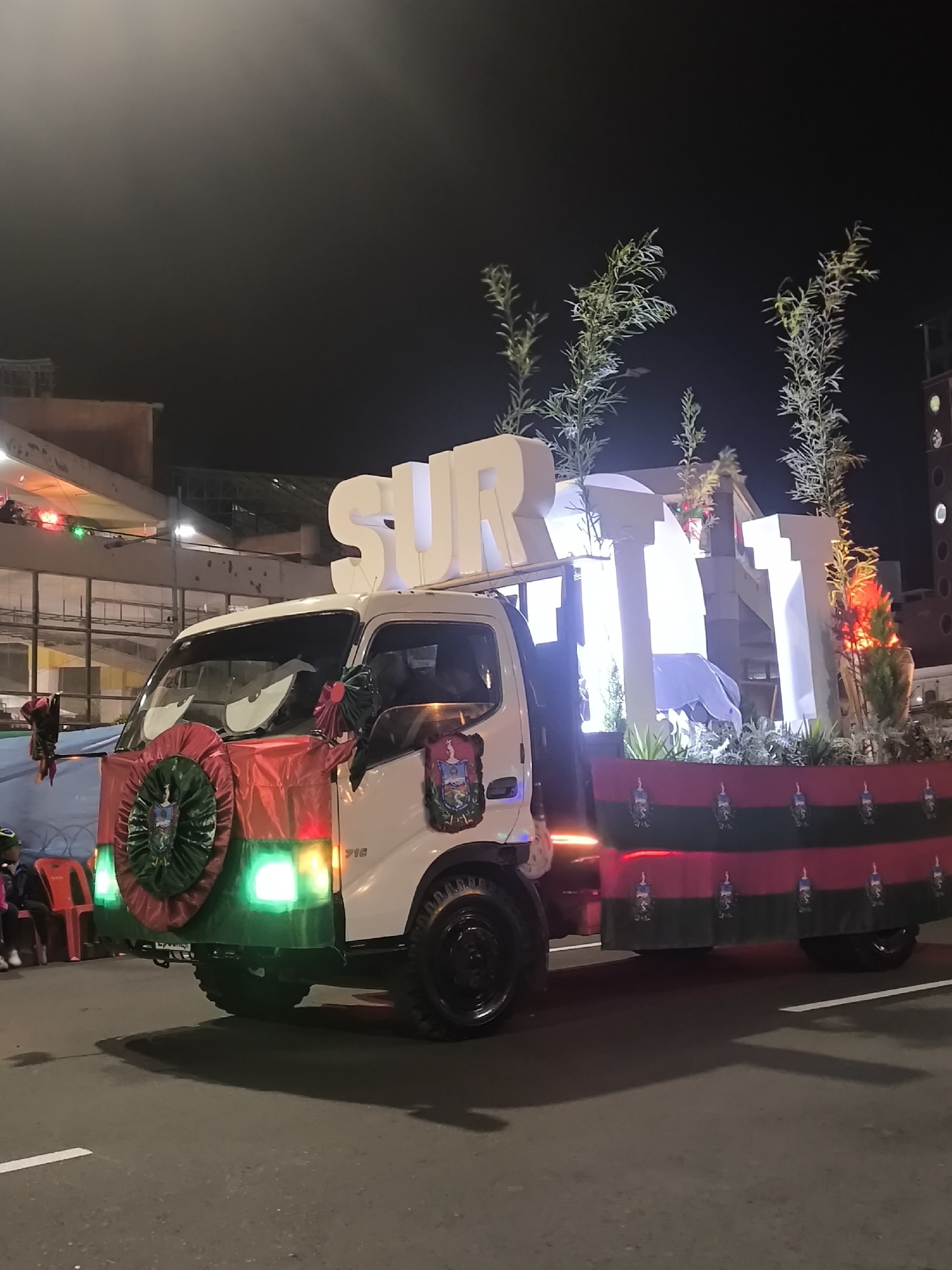 A white truck is decorated for a parade at night on a street in La Paz, Bolivia. The truck has large, white, illuminated letters spelling "SUR" on its roof. The front of the truck is decorated with a green and red circular emblem with a coat of arms, and large, cartoonish eyes are affixed to the windshield. The sides of the truck bed are draped with fabric featuring a repeating pattern of flags with a coat of arms. Green and red lights are visible on the front of the truck. Several people are visible in the background, sitting on red chairs. The environment appears to be an urban street at night, with buildings visible in the background.