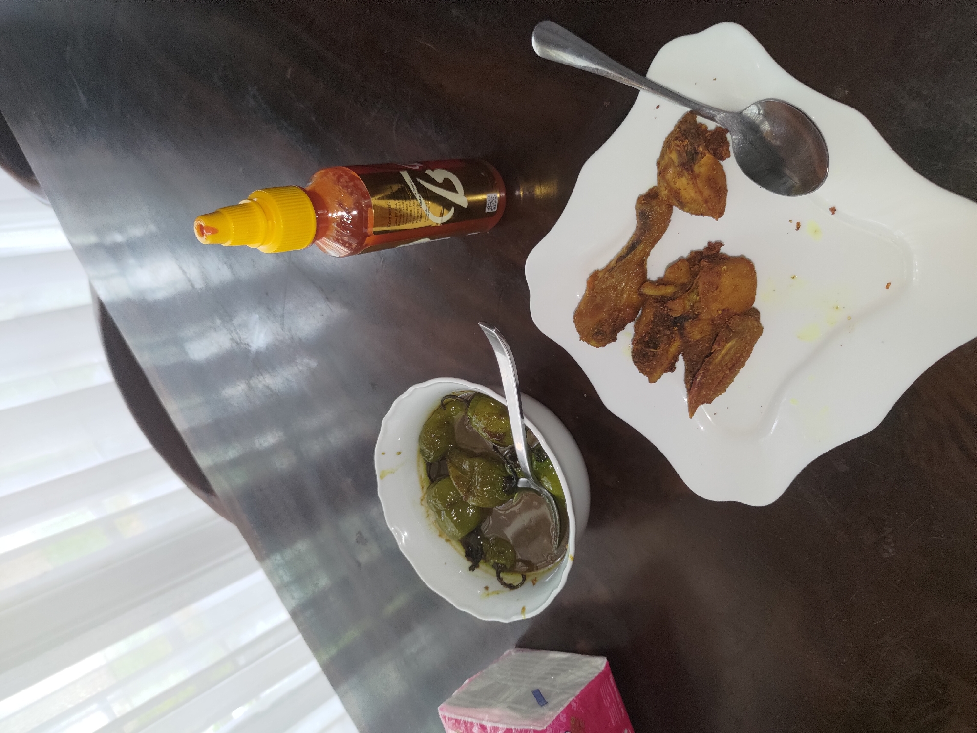 A close-up, high-angle shot shows a meal on a dark wooden table, likely in a home or restaurant setting in Depok, Indonesia. The table is partially covered by sheer white curtains in the background, allowing diffused natural light to enter the scene.
In the foreground, a white, wave-shaped plate holds several pieces of fried chicken. To the left of the plate is a white bowl containing green chilies in a brownish sauce, with a spoon resting inside. Next to the bowl is a pink and white box of tissues. To the right of the chicken plate, a metal spoon is placed. A red and yellow squeeze bottle of hot sauce stands upright behind the food. The overall impression is a casual meal setting.