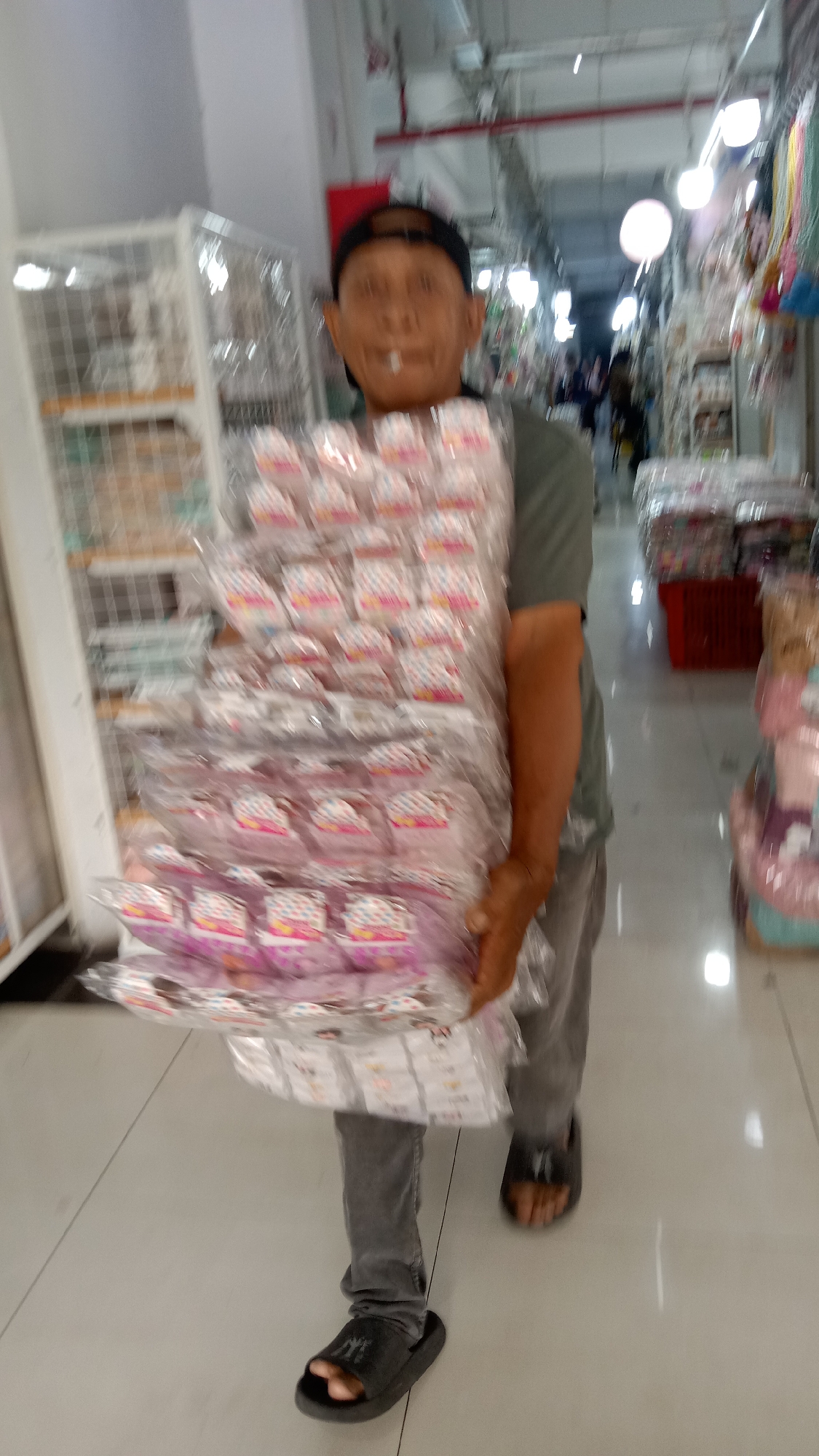 A full-shot, eye-level, slightly blurry, outdoor medium shot shows a person walking in a busy commercial street with stalls selling various goods in West Jakarta, Indonesia. The person, wearing a dark baseball cap, a gray t-shirt, and gray pants, is holding a large stack of individually wrapped items. The items appear to be small, possibly snacks or novelty items, packaged in clear plastic with colorful labels. The person is wearing black sandals. The background reveals a vibrant market environment with shelves stocked with merchandise and other shoppers blurred in motion, suggesting a lively commercial setting. The lighting suggests it might be daytime or a well-lit indoor market.