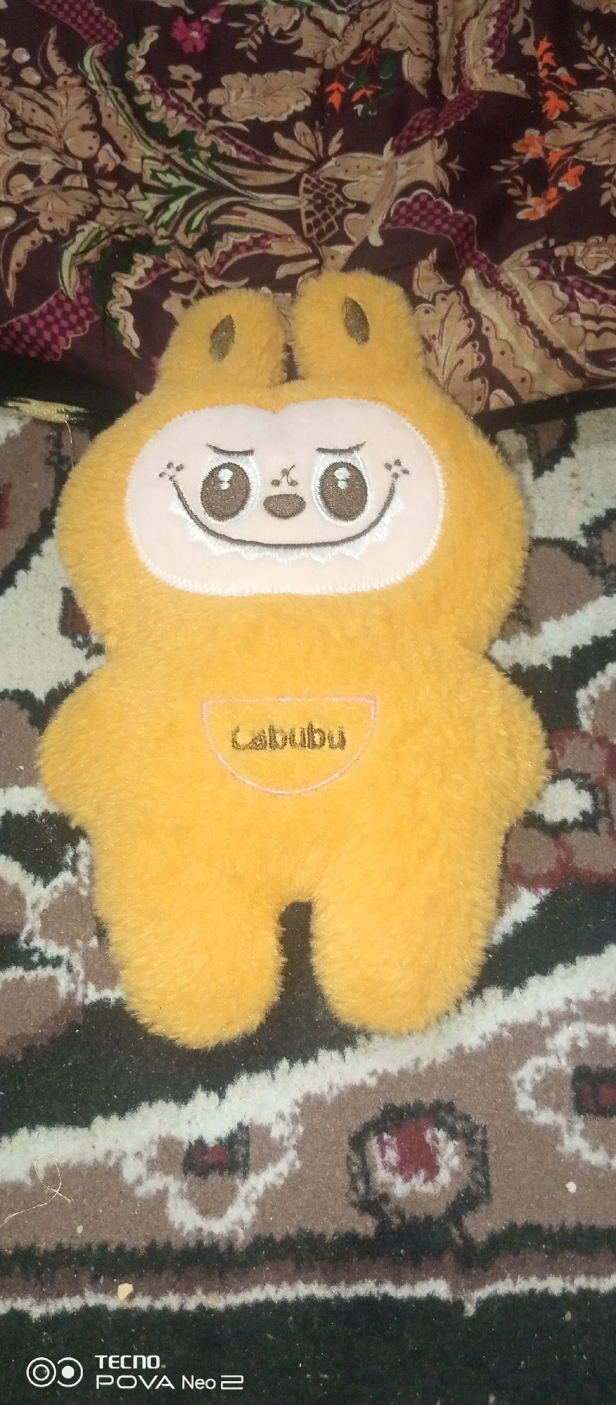 The image shows a close-up, top-down view of a fluffy, yellow stuffed toy on a patterned carpet. The toy has large, round eyes with black pupils, a wide, smiling mouth with a white, jagged edge, and two small, rounded ears at the top. On its chest, a pinkish-white outline encloses the word "Cabubu" in dark lettering. The carpet is a complex pattern of brown, black, white, and red, with geometric and floral elements. In the bottom right corner, a "TECNO POVA Neo 2" logo is visible. The scene does not provide any specific context or location within Tahmach, Uzbekistan.