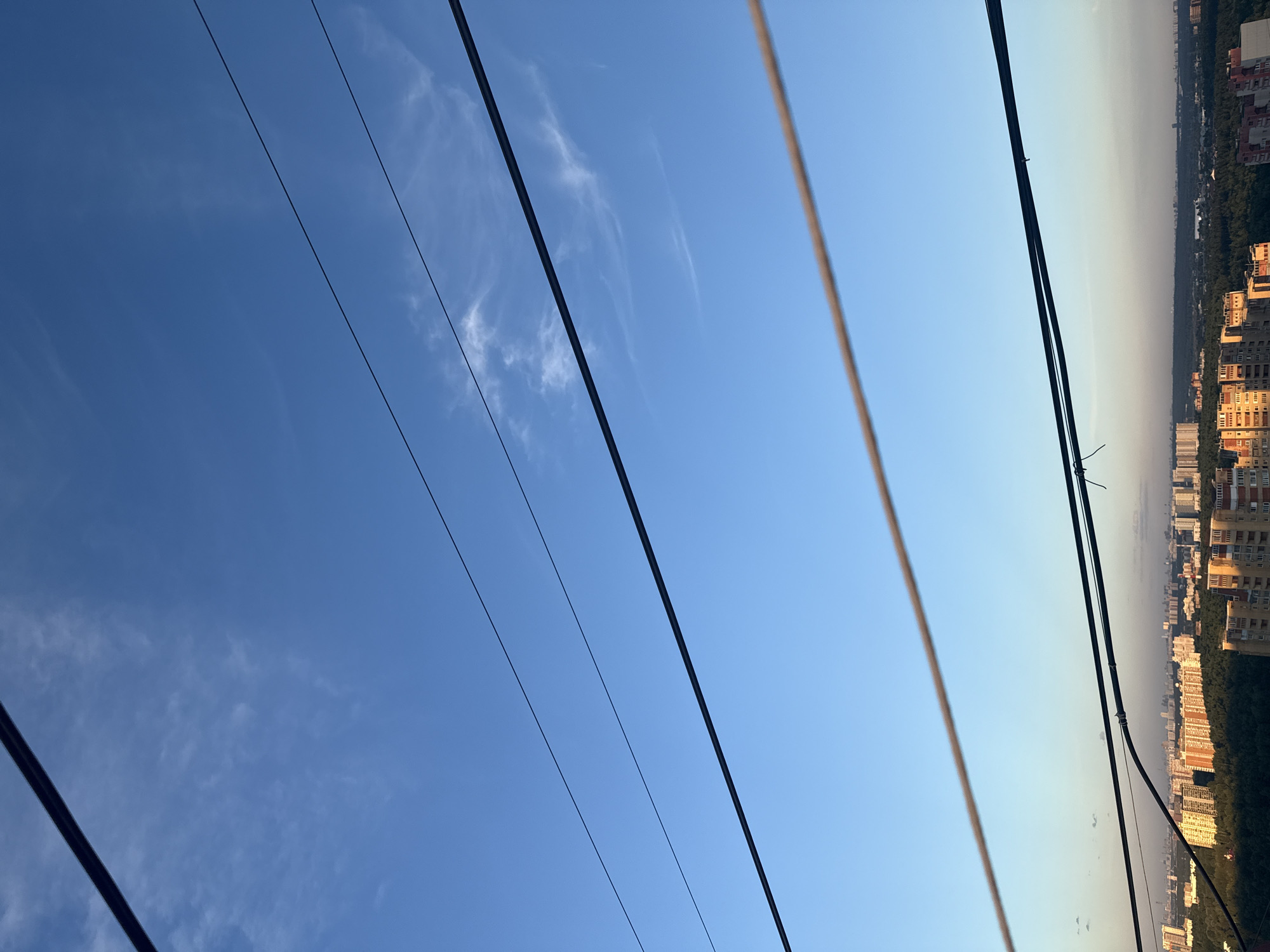 The image displays an outdoor scene with a clear blue sky filling the majority of the frame. Several dark, parallel wires run diagonally across the sky, suggesting overhead power lines. In the distance, a cityscape is visible on the right side of the image, with numerous residential buildings and trees. The orientation of the image is rotated 90 degrees counter-clockwise. The lighting suggests it is either early morning or late afternoon, with the sun casting a warm glow on the distant buildings. The weather appears clear with a few wispy clouds scattered in the sky. The scene is set in Khimki, Russia, as indicated by the location context. There is no visible text in the image.