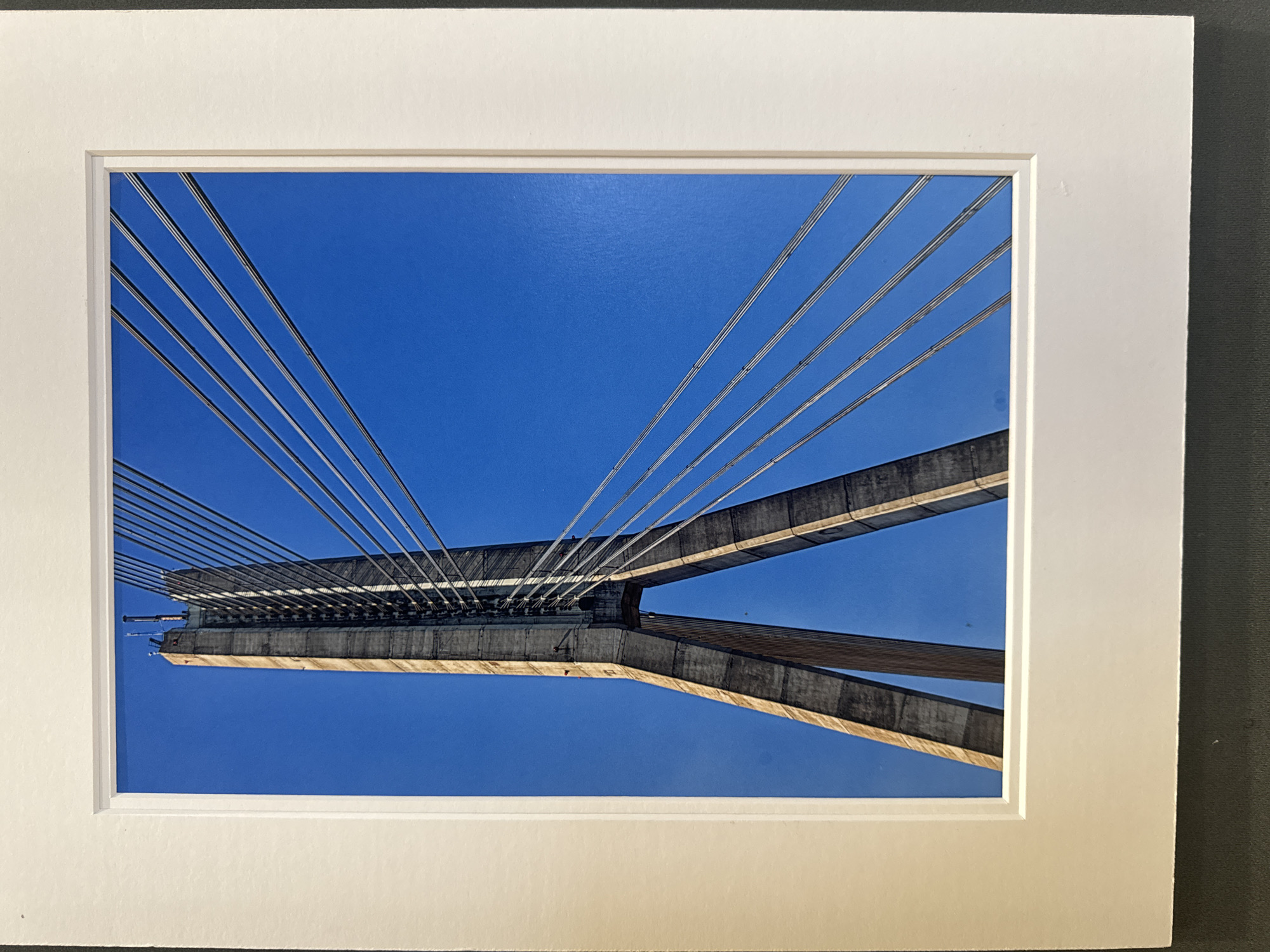 A low-angle photograph captures the imposing concrete pylons and steel cables of a suspension bridge against a vibrant, clear blue sky. The bridge pylons are tall and angular, tapering as they rise, with visible striations and structural elements. A multitude of parallel steel cables fan out from the top of the pylons, stretching diagonally across the frame and suggesting the immense scale of the structure. The lighting indicates a sunny day, casting sharp shadows and highlighting the textures of the concrete and metal. The image is framed by a white mat. This is a photograph of the Rose FitzGerald Kennedy Bridge in the city of Kilkenny, Ireland.