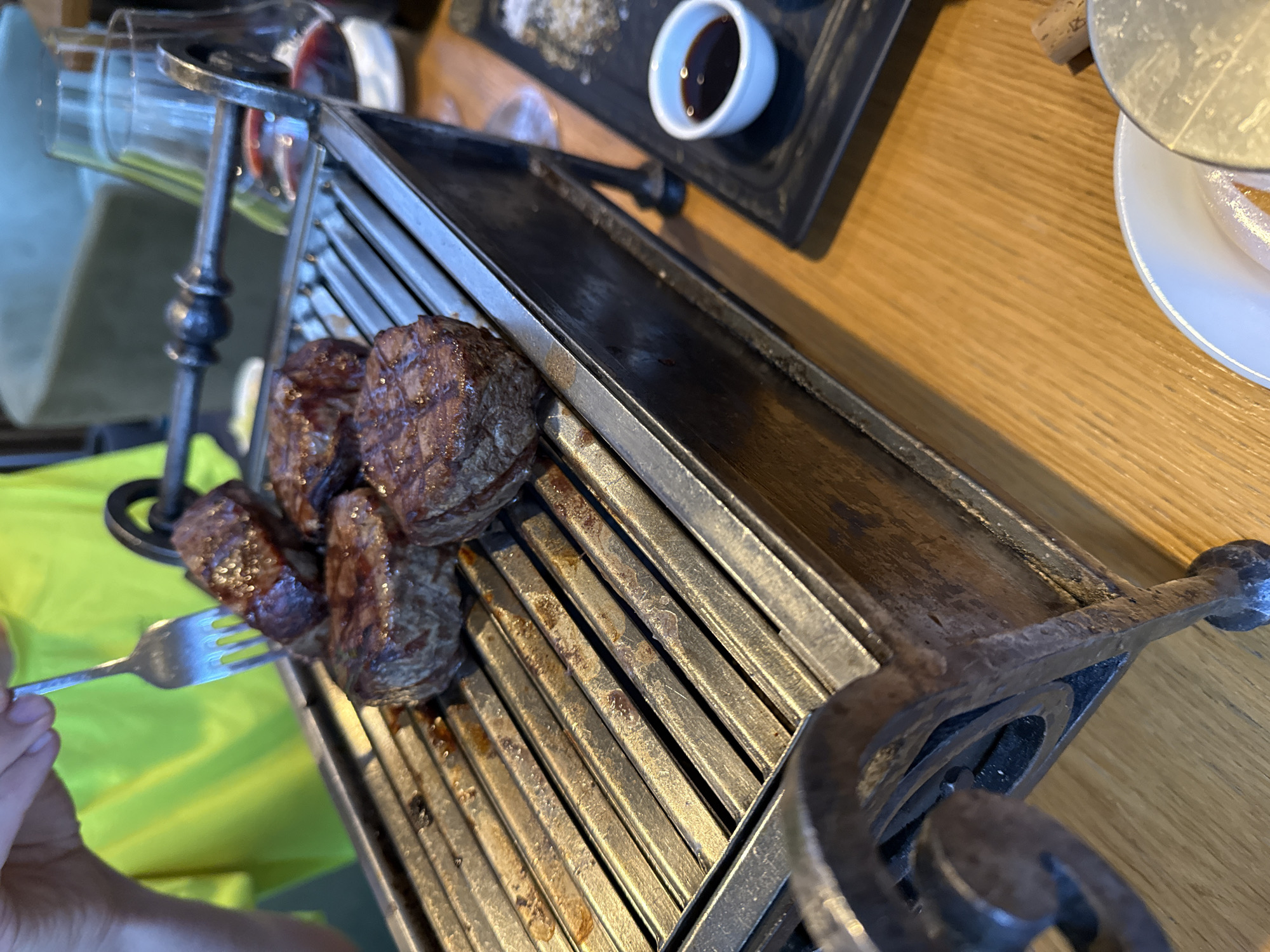 A medium, angled shot depicts a portion of a wooden table setting. On the table, a cast-iron grill-like platter holds four pieces of cooked meat. A silver fork is piercing one of the meat pieces. To the upper right, there are a small white plate and a small black tray containing a small white bowl with dark liquid. The scene appears to be in a restaurant. No specific location in Moscow, Russia, can be determined from the image alone.