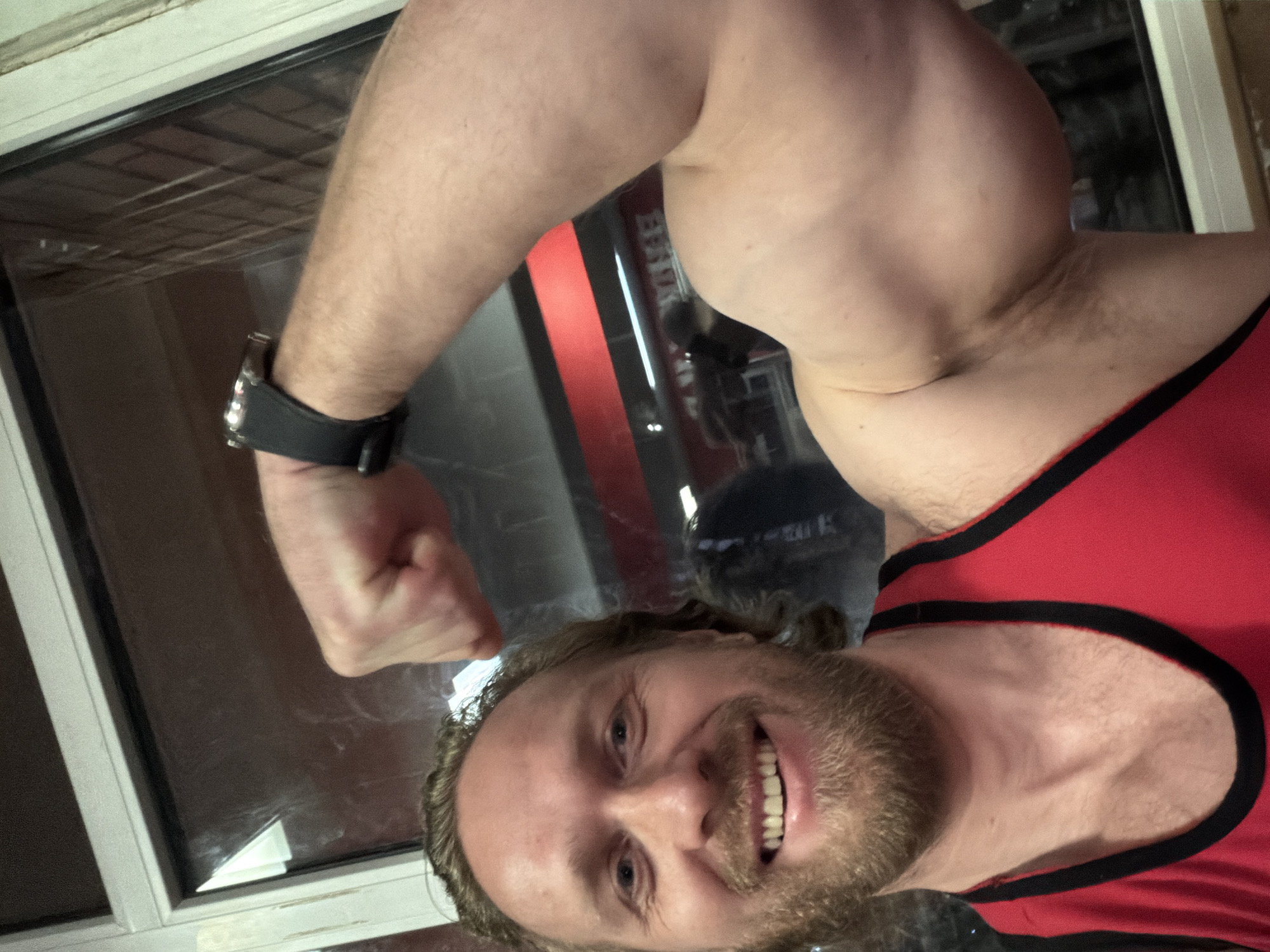 A male individual, appearing to be in his 30s or early 40s, with light skin, wavy light brown hair, and a full light brown beard, is depicted in a chest-up selfie. He is smiling broadly, revealing white teeth. The subject is wearing a red tank top with black trim. His right arm is flexed, displaying a prominent bicep. A black-strapped watch with a silver-colored casing is visible on his left wrist. The background features a window or reflective surface revealing an interior setting, likely a gym, with a horizontal red stripe on a wall and indistinct equipment visible in the reflection. The scene is located in the city of Saint Petersburg, Russia.