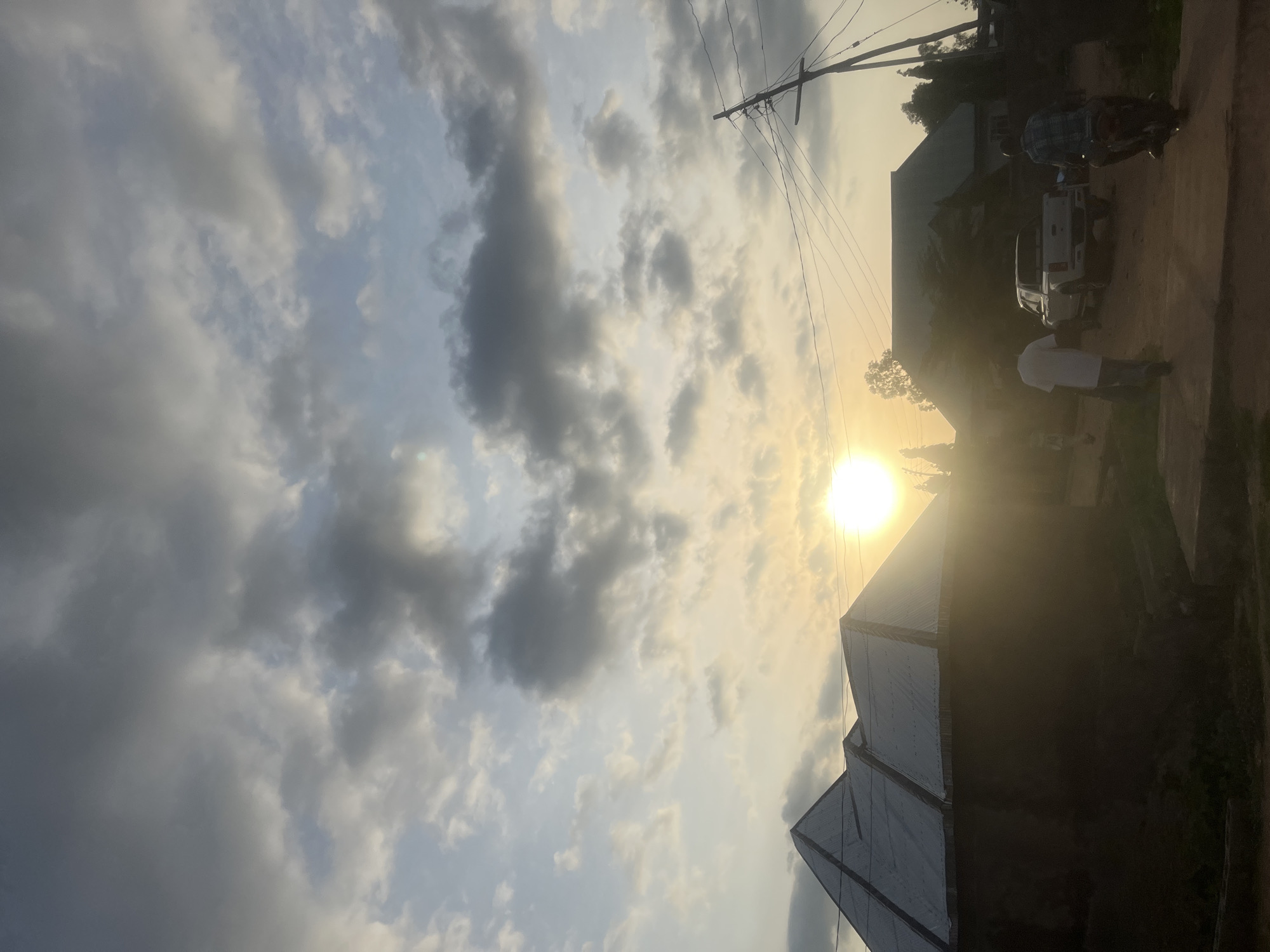 The image depicts a sunset scene in Jalingo, Nigeria. The sky is filled with a mix of grey and white clouds, with the sun prominently visible and casting a warm glow. In the foreground, a building with a corrugated metal roof is silhouetted against the light. Further down the road, a white pickup truck is parked, and two people are walking. On the right side of the frame, a person is riding a motorcycle. Utility poles with overhead wires are also visible, suggesting the presence of infrastructure in the area. The overall impression is of a rural or semi-urban setting at dusk.