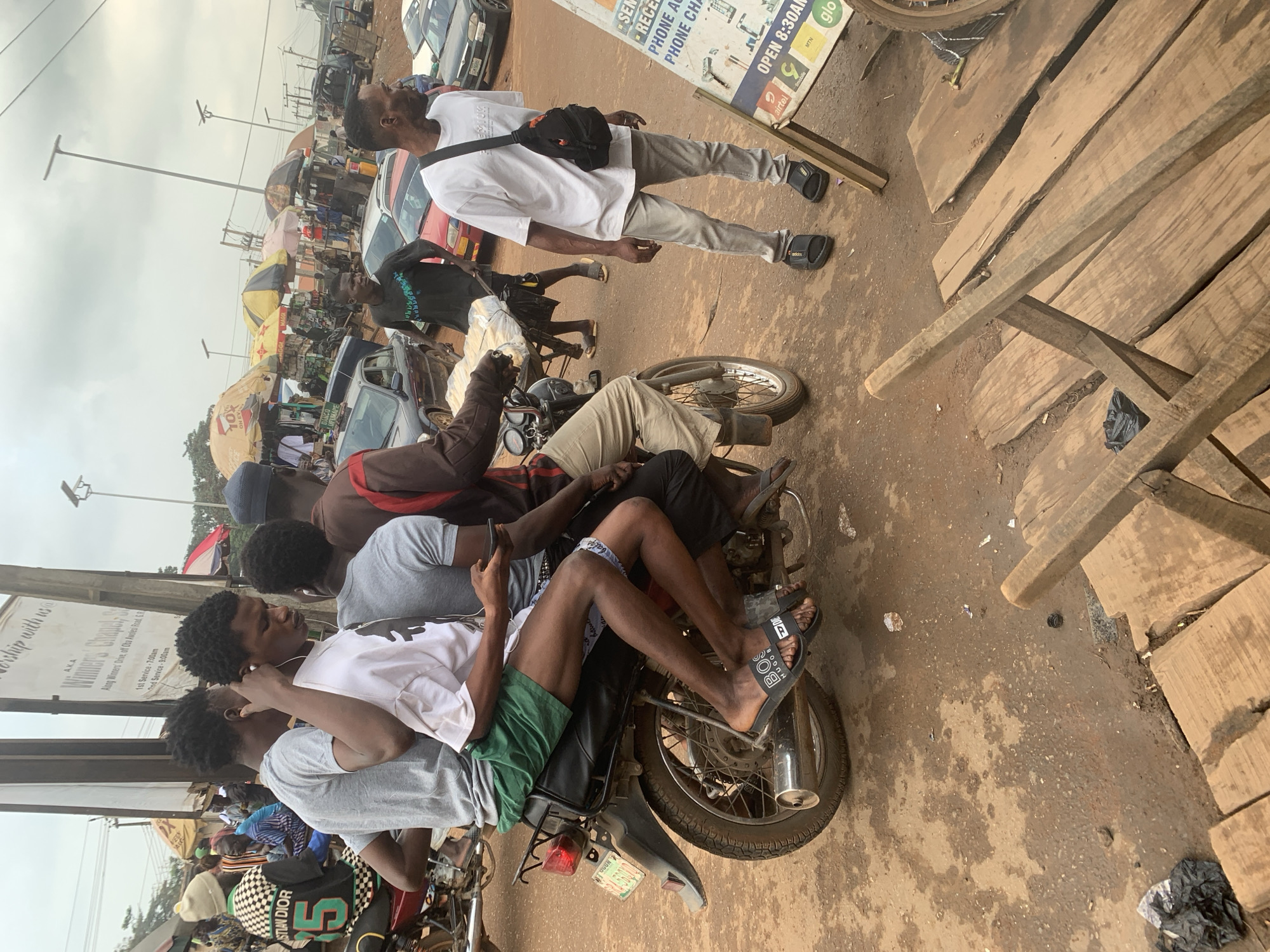 The image depicts a busy street scene in Shagamu, Nigeria, likely in a marketplace area. In the foreground, four young men are seated on a motorcycle. The driver, wearing a white t-shirt and grey pants, is holding a phone. Behind him, two men are also on the motorcycle, one in a grey t-shirt and green shorts, and another in a brown and red shirt. The fourth man is partially obscured.
The environment is a dirt road with scattered debris. To the left, a sign with text in black and white is visible, partially obscured by a motorcycle and the individuals. In the background, numerous market stalls with colorful umbrellas are set up, displaying various goods. Several cars are parked or driving on the road, and more people are milling about. The overall atmosphere suggests a lively commercial hub.