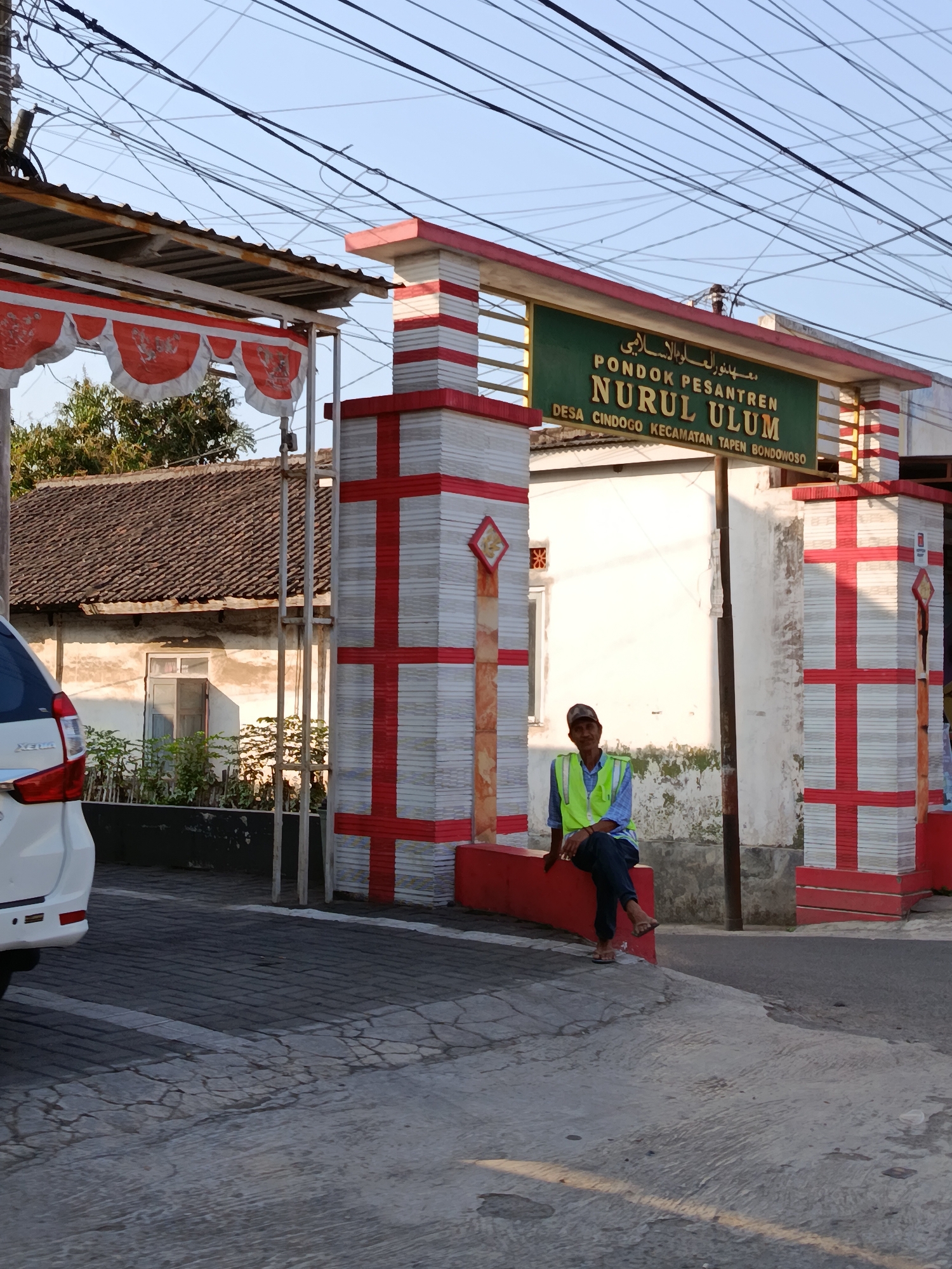 The image shows the entrance to the Nurul Ulum Islamic boarding school (Pondok Pesantren) located in Cindogo Village, Tapen District, Bondowoso City, Indonesia. A man wearing a green safety vest and a blue plaid shirt is sitting on a red concrete barrier in front of one of the school's pillars. The pillar is decorated with red and white checkered patterns. To the left, a white car is partially visible, and beyond it is a building with a tiled roof and a window. The background features a clear sky with numerous electrical wires crisscrossing overhead. The overall scene depicts the exterior of an educational institution entrance in a semi-urban or rural setting.