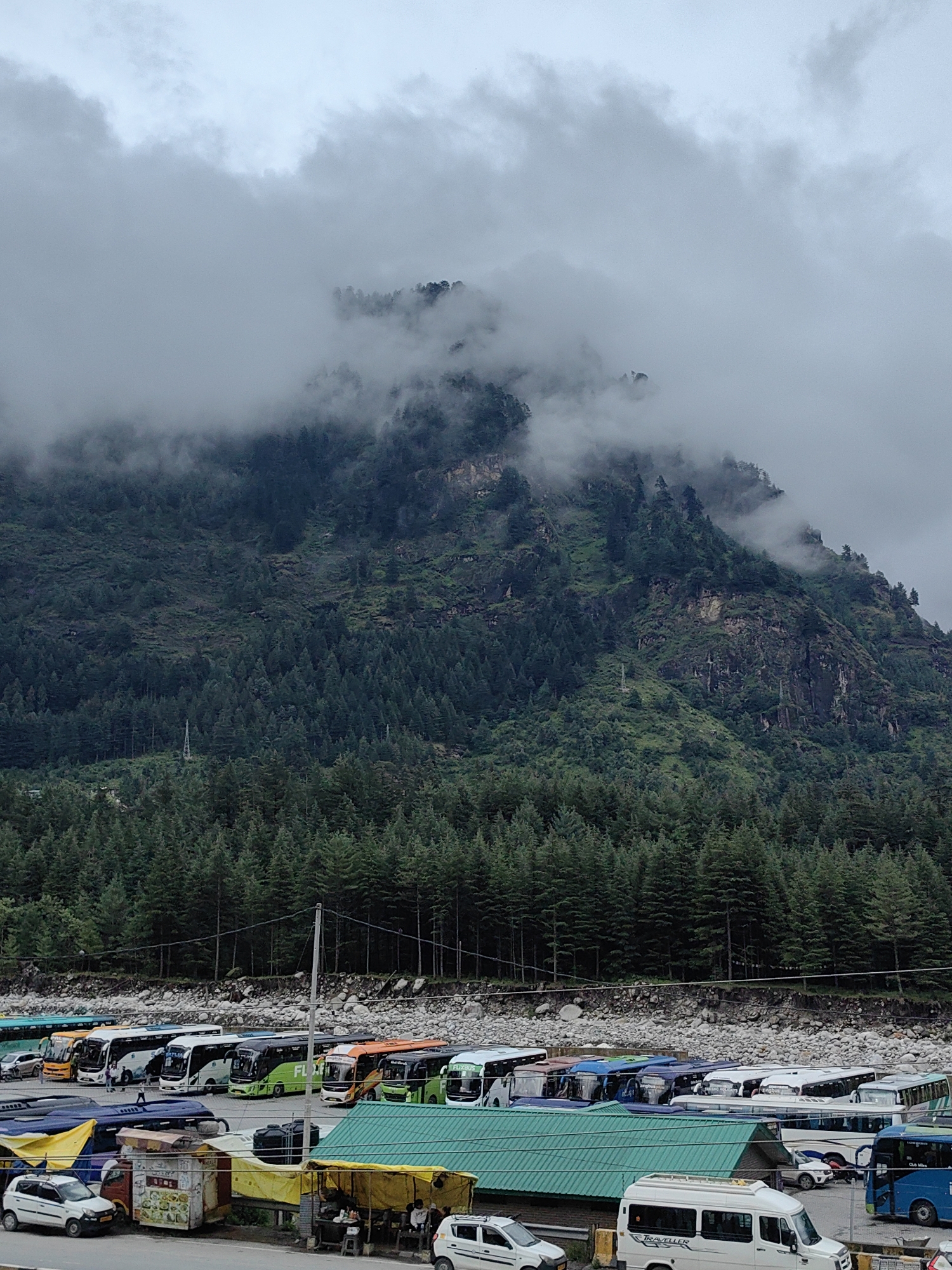 A cloudy, overcast day in Siumsa, India, reveals a parking lot filled with buses and other vehicles at the base of a mist-shrouded mountain covered in dense evergreen trees. In the foreground, a row of buses, predominantly white, green, and orange, are parked side-by-side. Some buses have text such as "FLYBUS" and "INDIGO" visible on their sides. Several smaller white cars and a white van are also present. A few people are visible near the buses and in the background, near small food stalls. The mountain behind the parking lot is heavily forested with tall evergreen trees, and the upper reaches are obscured by thick, white clouds and mist, giving the scene a somewhat ethereal quality. The terrain is rocky and appears to be on the side of a valley or mountain pass. The overall atmosphere is damp and cool, typical of a mountainous region with cloud cover.