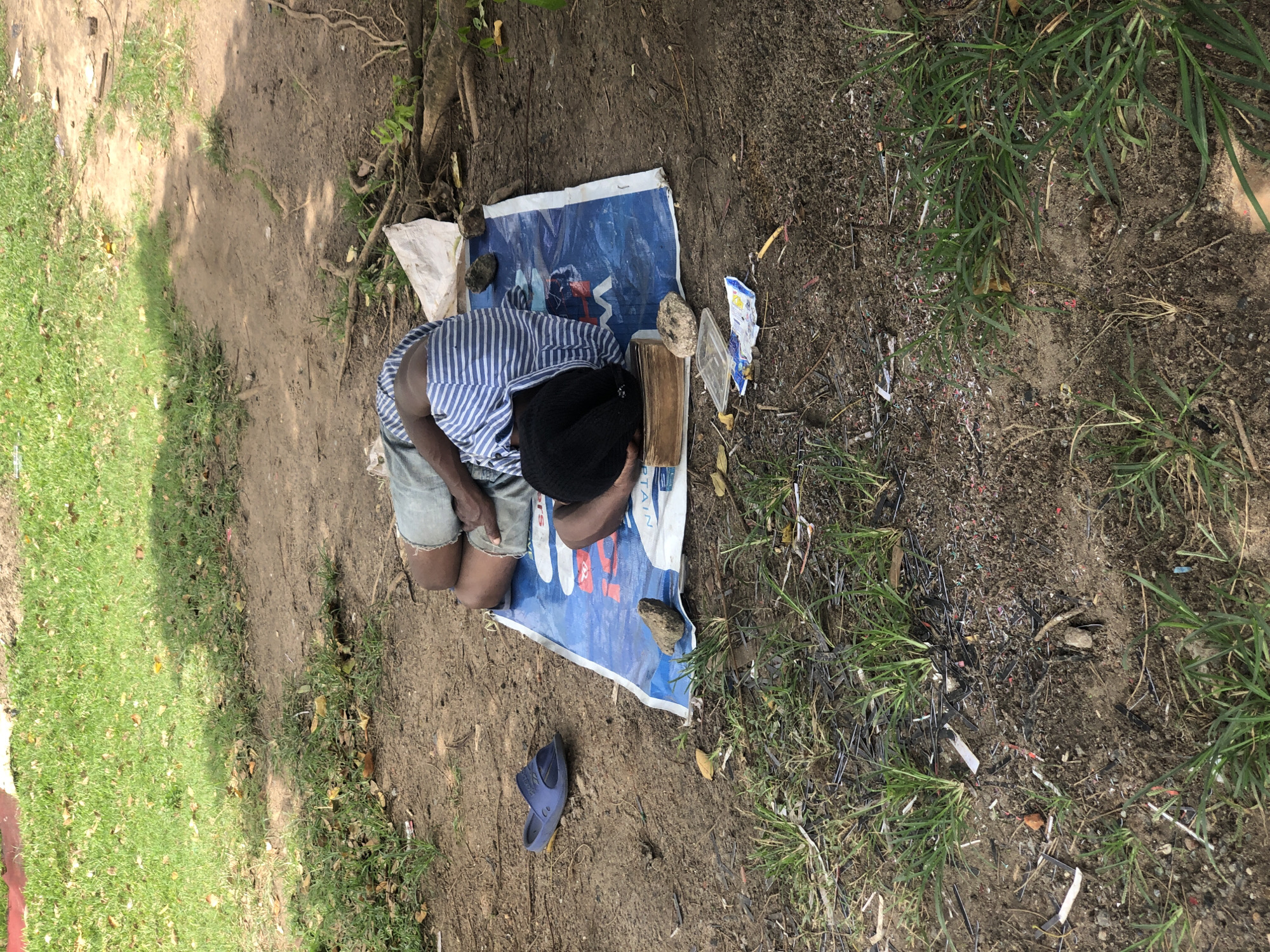 A person is sleeping on a blue tarp on the ground in a wooded area. The person is wearing a striped shirt and shorts. There is a blue sandal to the left of the tarp and a pile of books under the person's head. The ground is dirt and there is grass and leaves around the area. The location appears to be a park or a natural area within the city of Ikeja, Nigeria.