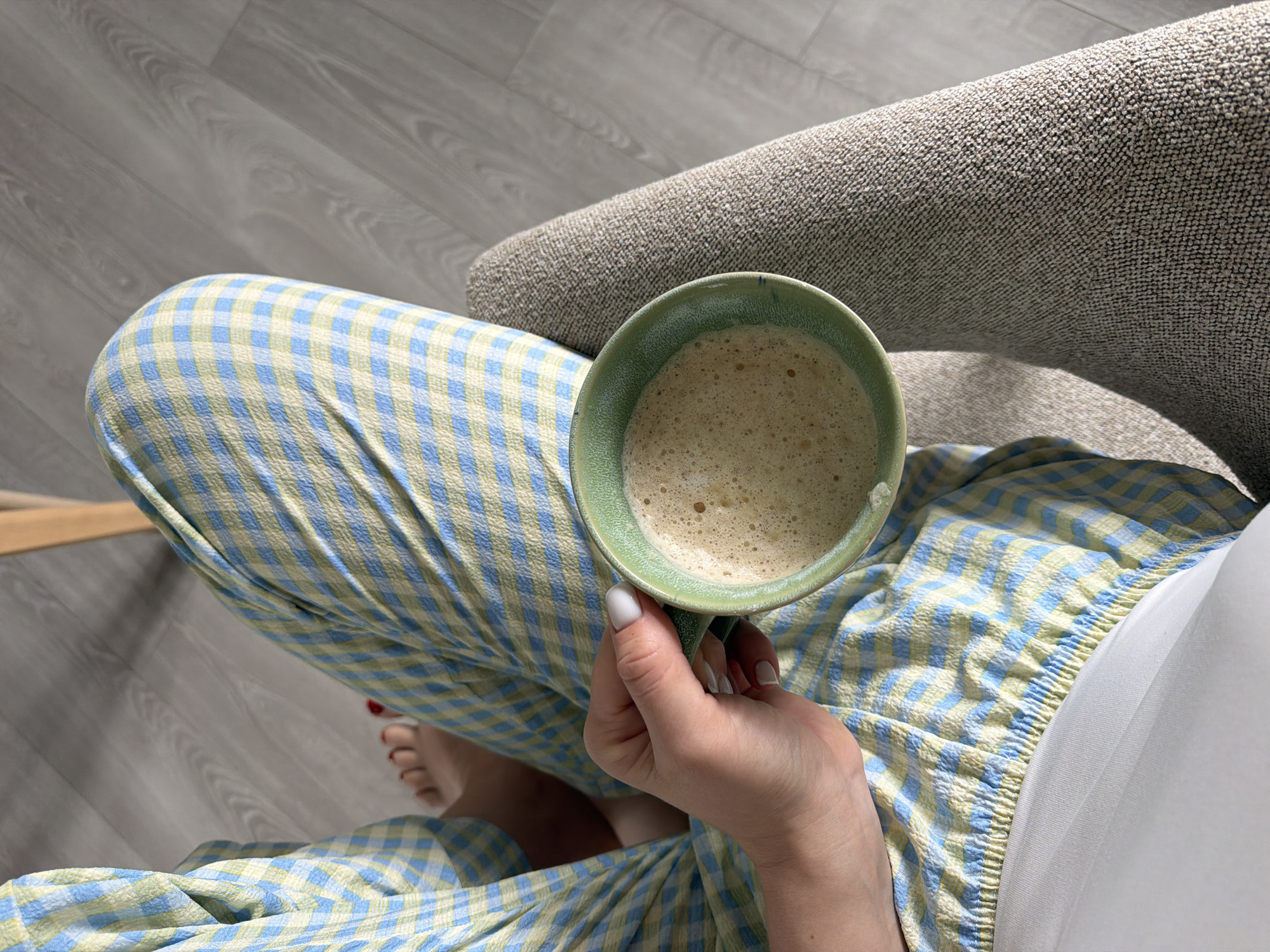 An overhead, close-up shot shows a person sitting in a chair, holding a green mug filled with a frothy beverage. The person is wearing blue and yellow checkered pajama pants and has a white painted fingernail. Their feet, with red painted toenails, are visible in the background. The floor appears to be light wood. The overall scene suggests a casual, domestic setting, likely a home. There is no information in the image that would indicate a location in Dreiliņi, Latvia.