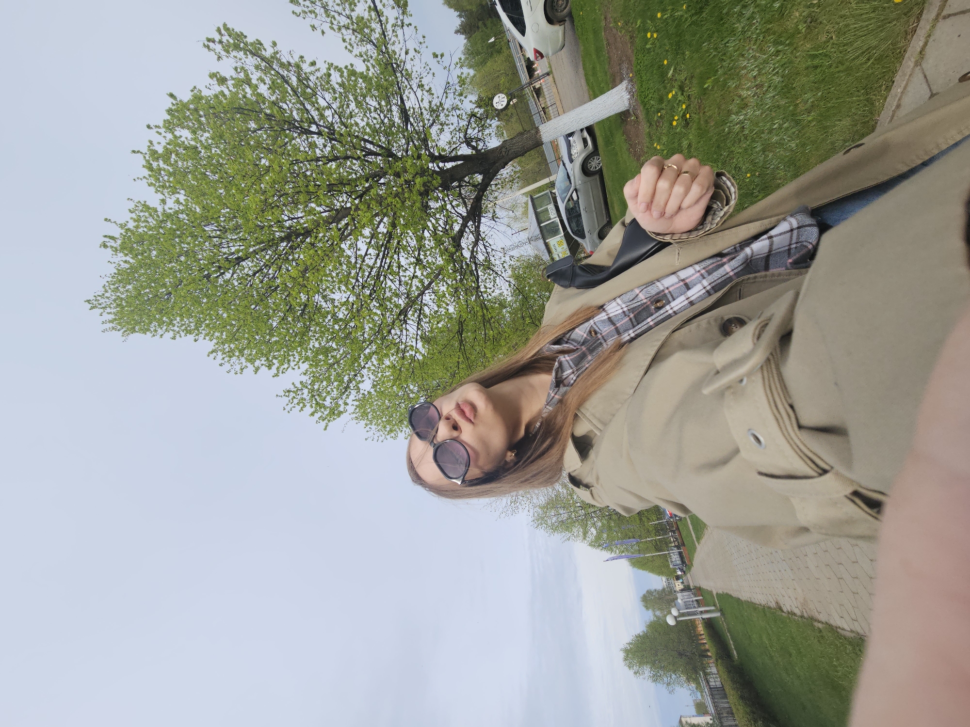A young woman in sunglasses and a tan coat stands outdoors. She is wearing a plaid shirt under the coat. She holds her fist near her face, showing two gold rings on her fingers. Behind her is a tree with green leaves. There is grass and a sidewalk to the right. Cars and a building are visible further back. The scene is in the city of Chaikovsky, Russia.