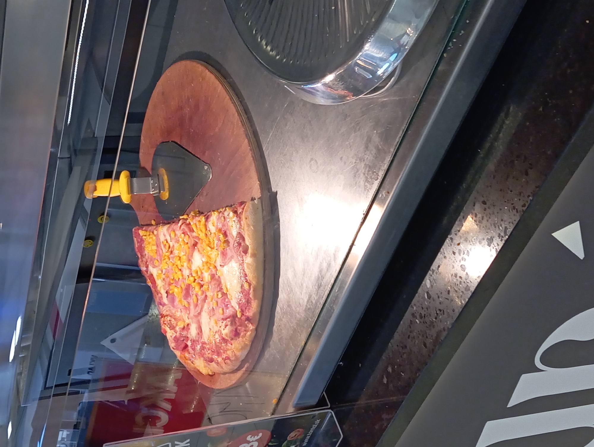 This image captures a food display case within a commercial establishment, specifically a Lanta fast-food location in Nitra, Slovakia, indicated by the partial "www.lanta.sk" text on a visible sign. The central subject is approximately half of a large pizza, resting on a circular wooden pizza board. The pizza is topped with sliced ham, abundant yellow corn kernels, and a scattering of white flakes, possibly cheese or seasoning, over a light-colored base and golden-brown crust. A metallic pizza server with a bright yellow handle is positioned on the wooden board, adjacent to the pizza.
The pizza and serving utensil are enclosed within a clear glass display counter. The counter's base is a brushed metallic surface, showing minor scratches and reflections. Above the pizza, additional glass shelving and metallic structures are visible. Reflections on the display glass include the inverted red text "AKCIA" (Slovak for "SALE" or "PROMOTION") and indistinct interior elements, along with small yellow smiley-face decals on a horizontal bar in the background. Behind the display, a light blue wall or panel and a white, conical object are discernible.
To the left, a vertical clear acrylic sign is partially visible, displaying "3€", indicating a price in Euros, and the aforementioned "www.lanta.sk" address, affirming the location. The bottom portion of the image reveals a dark, speckled countertop, reflecting overhead light. No individuals are present in the scene, which depicts a static food display.