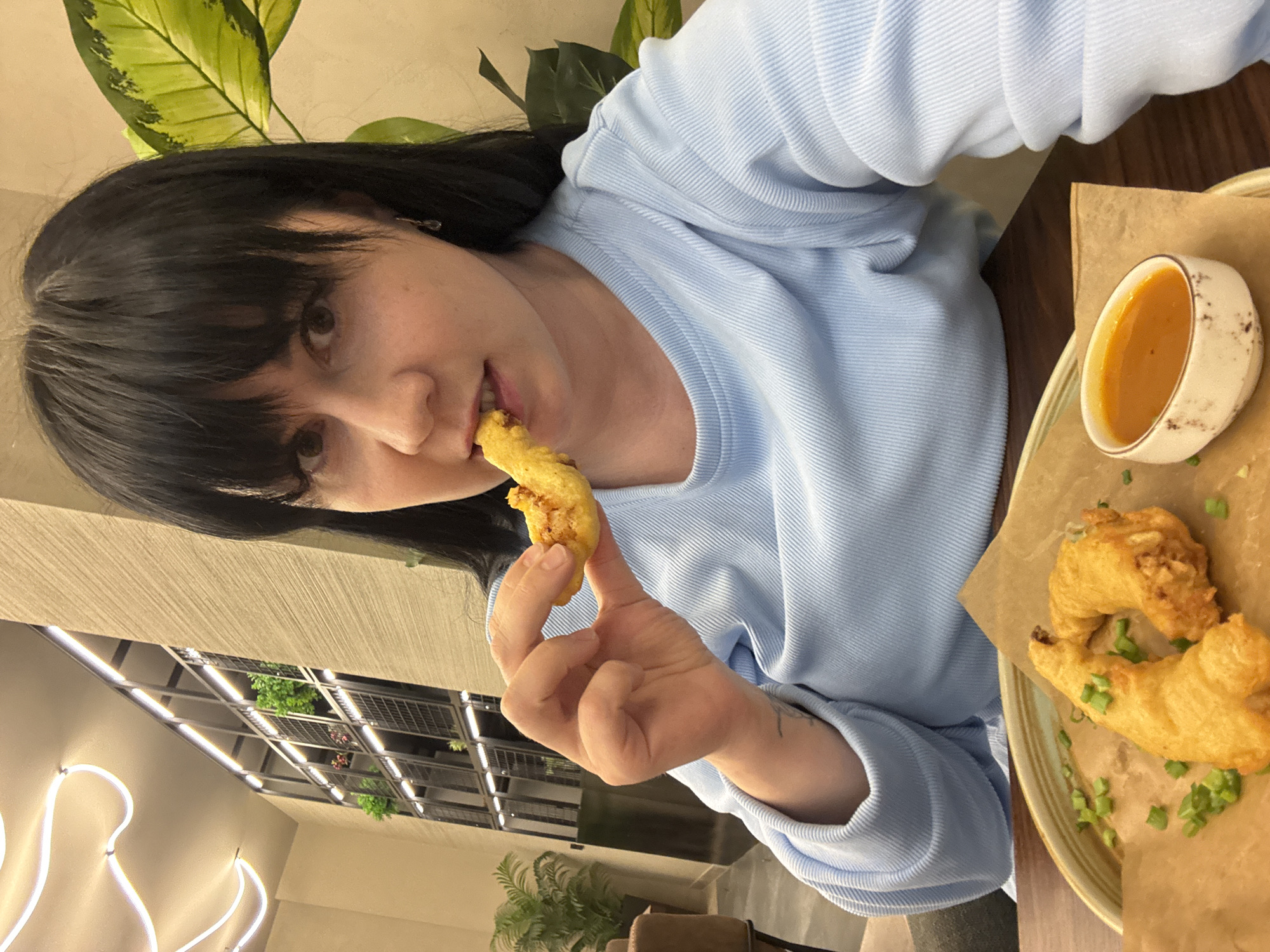 A medium, slightly angled, indoor shot shows a woman with dark hair and bangs, wearing a light blue, horizontally ribbed sweater, holding a fried food item to her mouth. She is sitting at a table with a plate of fried food and a small white bowl of sauce to her right. The food appears to be garnished with green onions. Behind her and to the left, a neon light fixture is visible on the wall, and through a window, a grid-like structure with greenery can be seen. A potted plant is also visible near her in the background. The restaurant’s location appears to be Izhevsk, Russia.