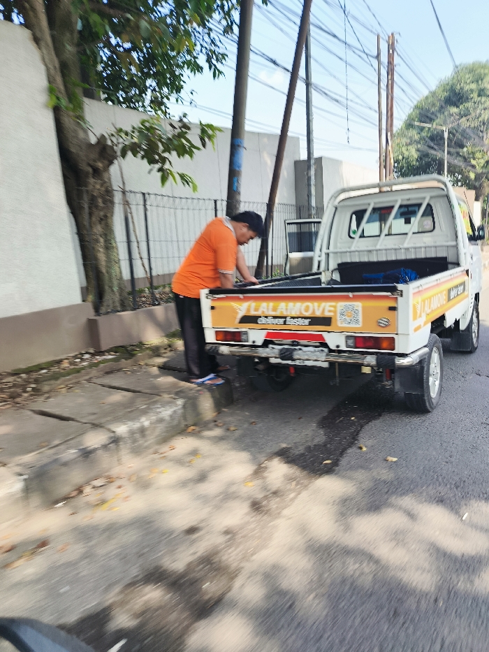 A white pickup truck with a yellow and orange "Lalamove" logo is parked on a street in Ngamprah, Indonesia, at the coordinates -6.867 latitude, 107.518 longitude. A man in an orange shirt is standing near the truck, possibly preparing to load or unload items. There is a tree to the left of the truck, and a concrete curb on the right side of the truck. The truck is partially obscured by a shadow from the tree. The street is paved, with some visible tire tracks and a dark liquid spilled on the pavement.