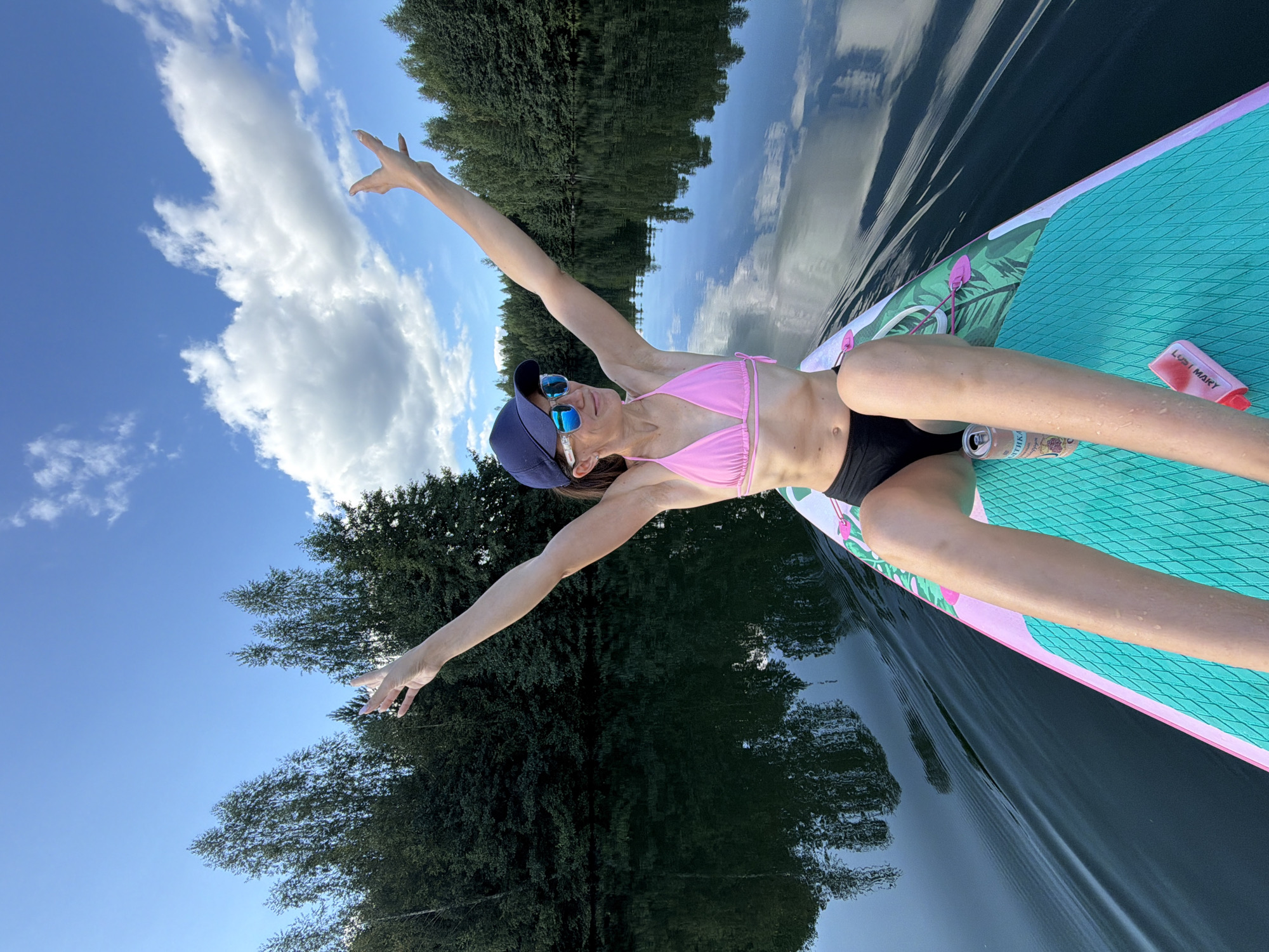 An image shows a woman paddleboarding on a calm lake surrounded by trees. The woman is wearing a pink bikini, black shorts, and a dark blue baseball cap. She has sunglasses on and her arms are raised in the air. The paddleboard is decorated with a tropical pattern. The sky is blue with some white clouds. The water is dark and reflects the trees and the sky. The scene appears to be in a natural setting, possibly near a forest. The location is identified as Chernushka, Russia.