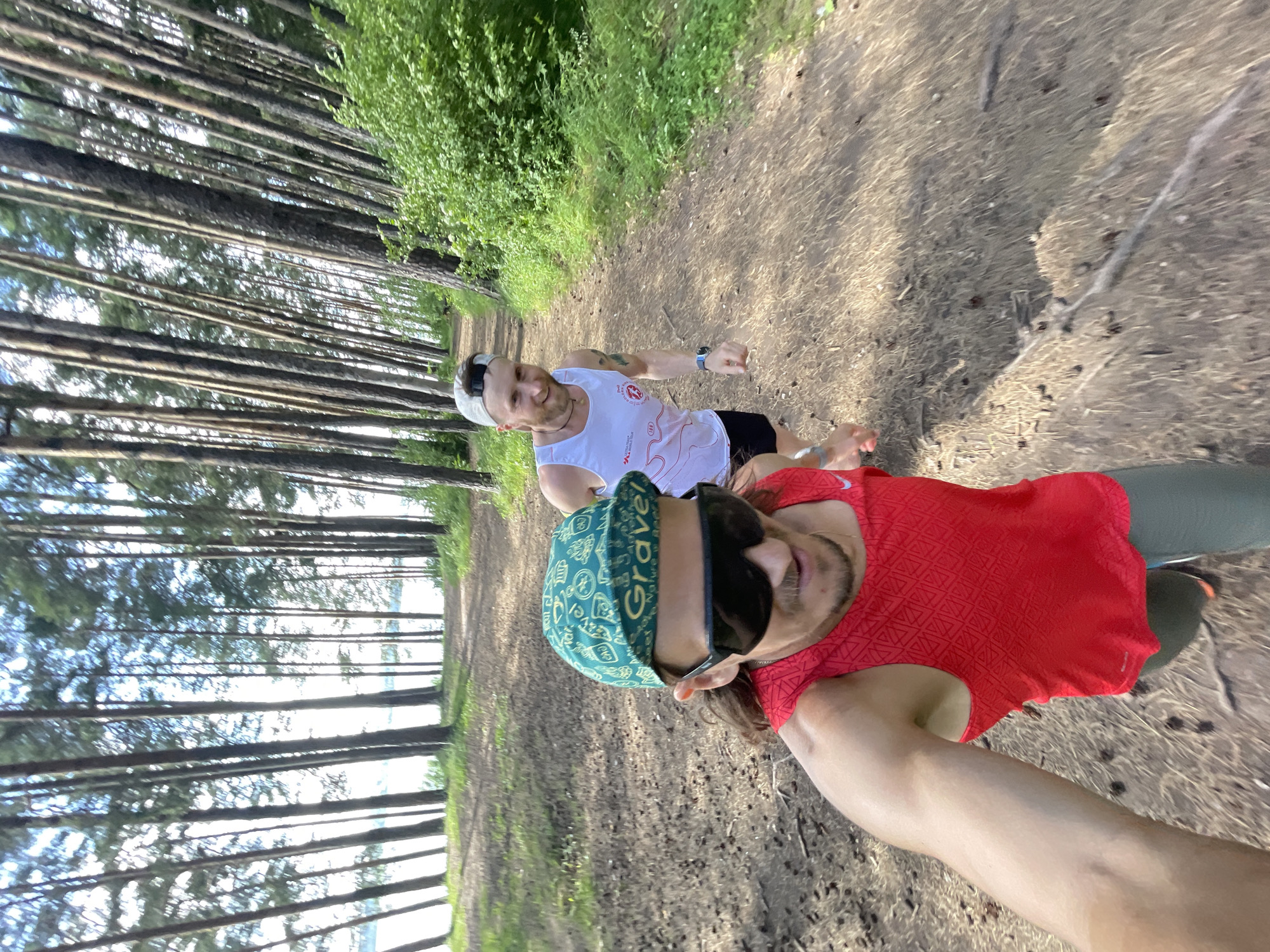 Two men are running on a dirt path in a forest. The man in the foreground is wearing a red tank top, green baseball cap, and sunglasses. He is holding the camera and appears to be taking a selfie. The man in the background is wearing a white tank top and a grey baseball cap. He is smiling and looking at the camera. The path is lined with tall pine trees and green bushes. The sun is shining through the trees, creating dappled light on the path. There is a body of water visible in the background, beyond the trees.