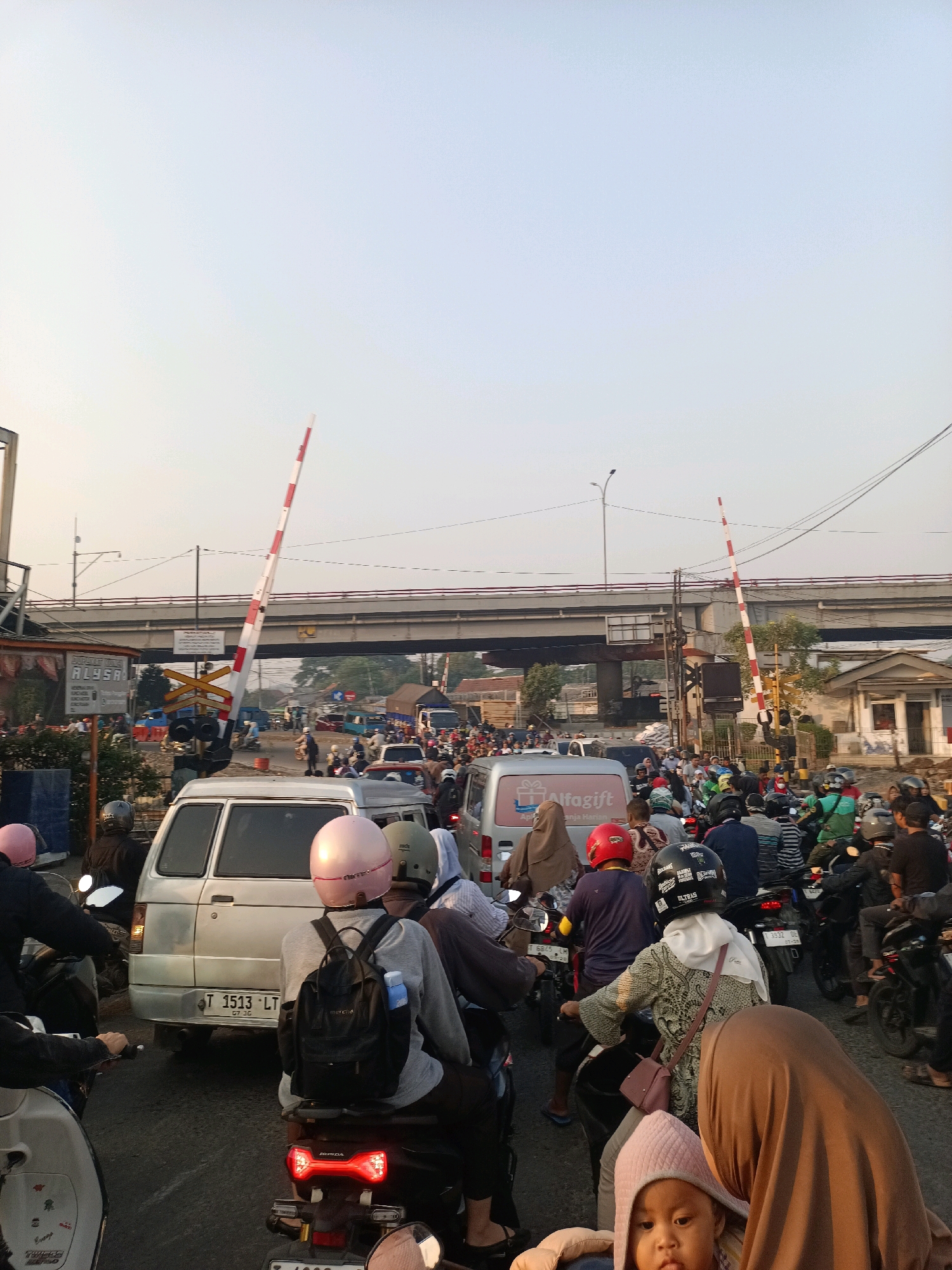The image depicts a busy traffic scene at a railway crossing in Karawang, Indonesia. A red and white striped barrier arm is lowered, indicating an approaching train. A large number of motorcycles and some cars are stopped, forming a traffic jam. Several people are visible riding motorcycles, many wearing helmets. The sky is clear with a pale yellow hue, suggesting it is either early morning or late afternoon. In the background, an overpass or bridge is visible, along with buildings and trees. The overall atmosphere is one of congestion and waiting.