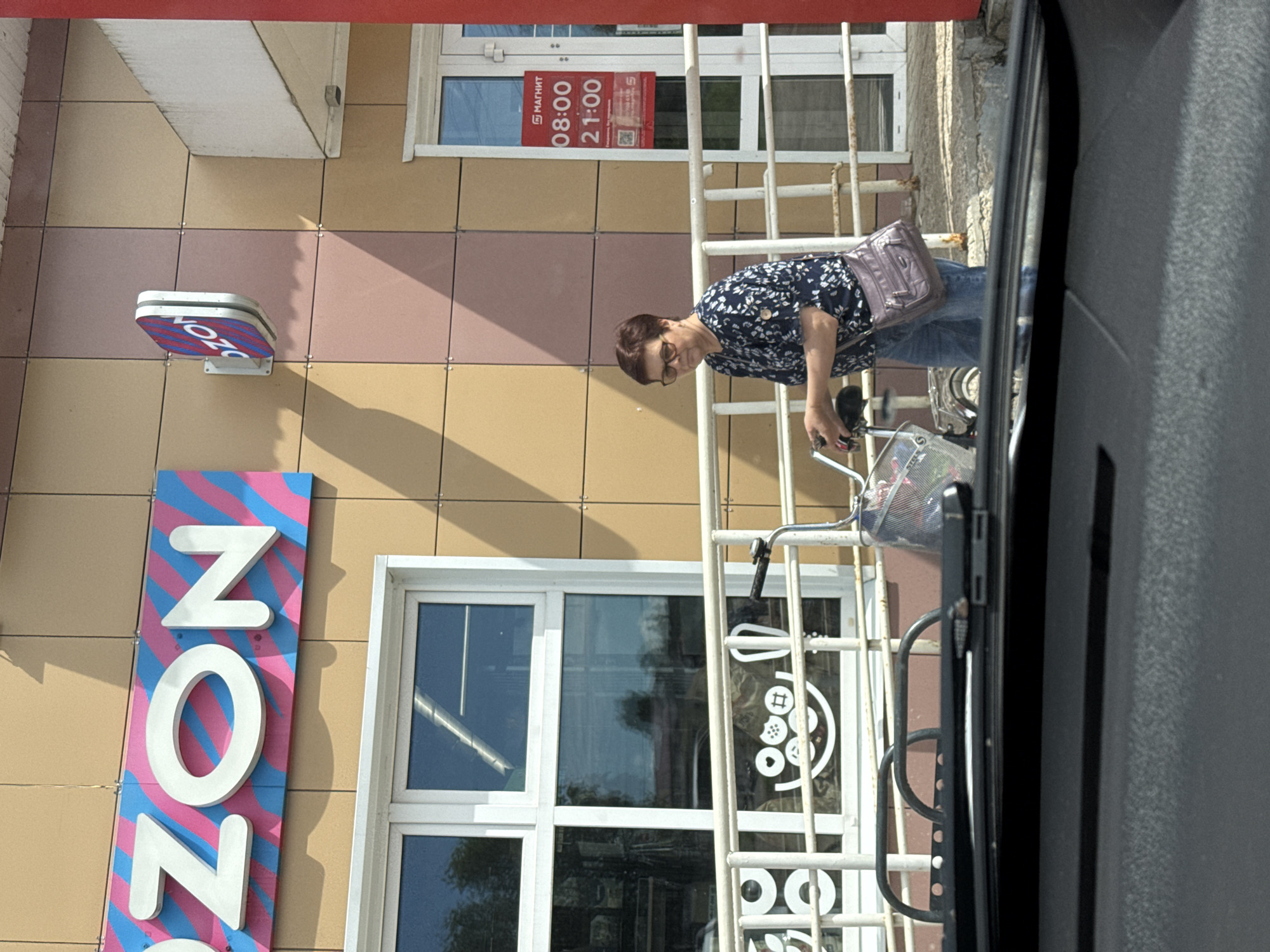 In Valday, Russia, a woman with short brown hair and glasses, wearing a dark blue floral print blouse and blue jeans, walks with a bicycle in front of a building. The building has a facade of alternating beige and pinkish-brown square tiles. A large sign with the letters "OZON" in white 3D lettering against a pink and blue striped background is mounted on the building. To the right of this sign is a smaller, oval-shaped sign also displaying the "OZON" logo. To the right of the woman, a red sign with white text indicates store hours: "08:00" and "21:00" with a "МАГНИТ" logo above. The woman is carrying a purple purse on her left shoulder. She is pushing a bicycle with a metal basket on the front, containing a pink plastic object and other items. A white metal railing with decorative circles and shapes is in the foreground, partially obscuring a window with a reflection of trees and sky. The scene appears to be captured from inside a vehicle, as indicated by the presence of a windshield wiper in the bottom of the frame.
